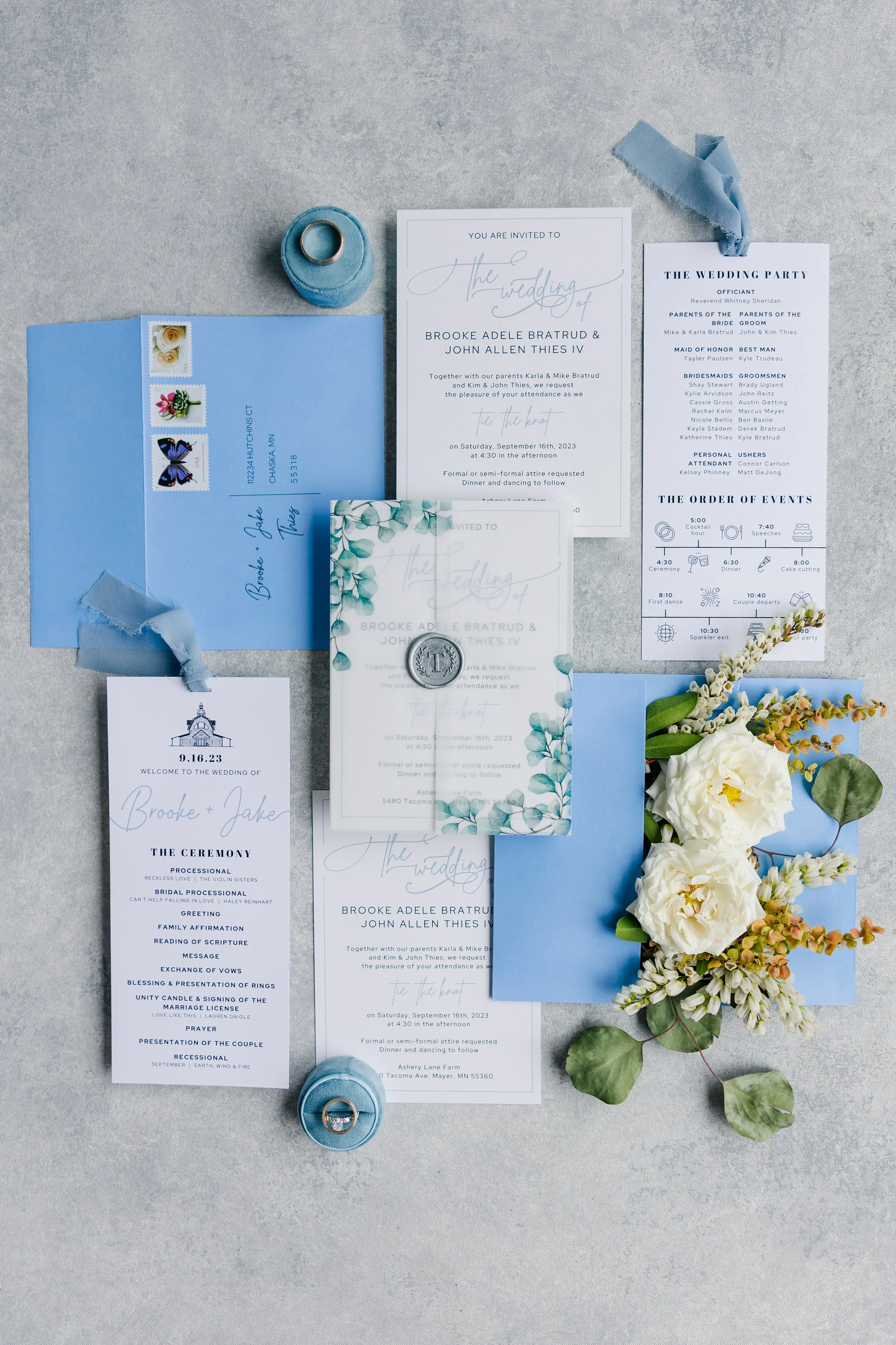 Wedding invitation suite with floral accents, blue envelopes, RSVP card, wedding program, and bouquet of white roses and greenery.