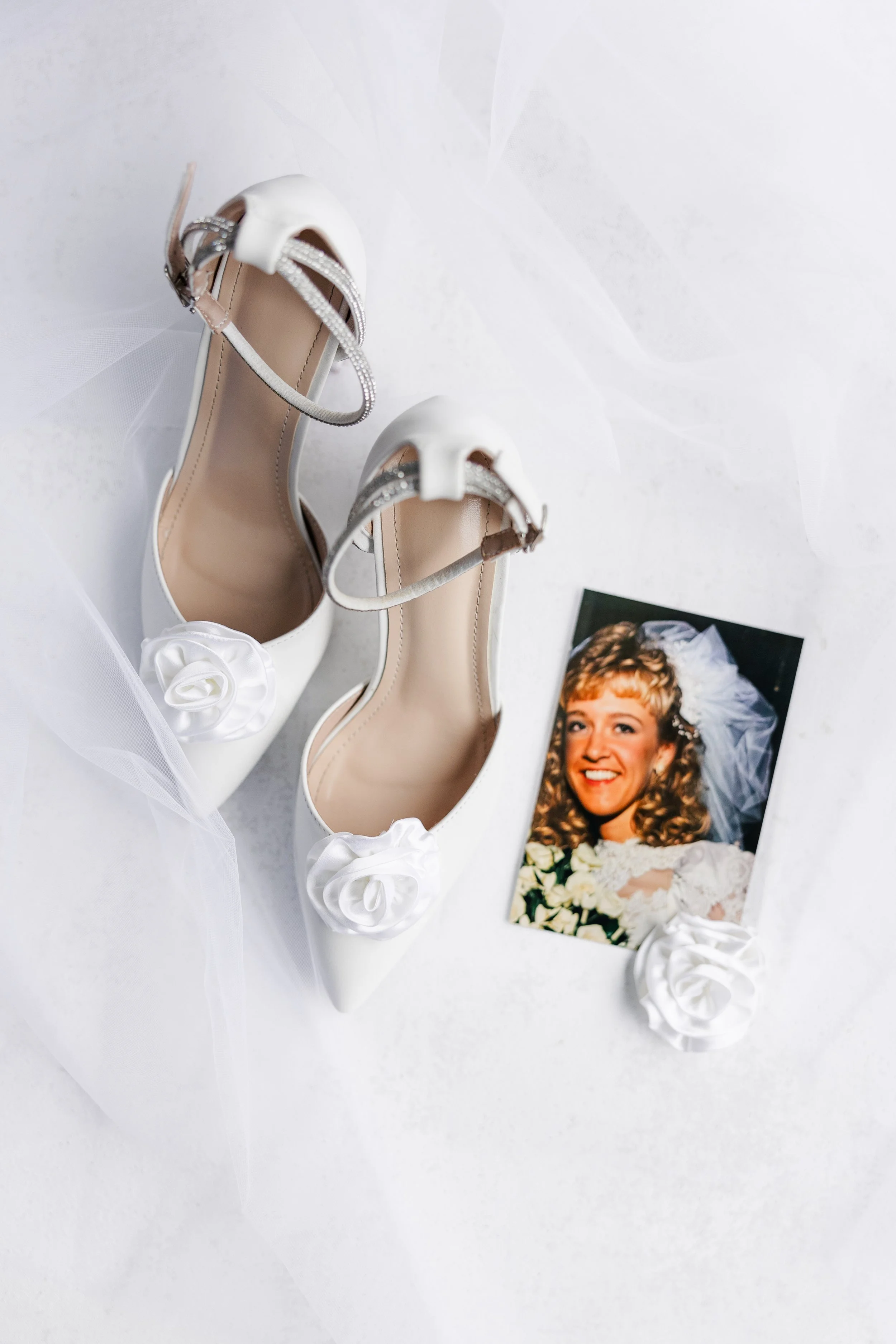 A pair of white wedding shoes with floral embellishments, a tulle veil, and a photograph of a smiling bride with short blonde curly hair, wearing a veil and holding a bouquet of white flowers.