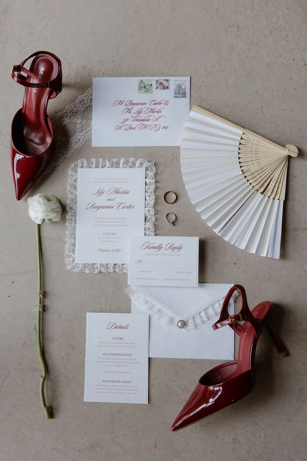 A wedding invitation suite with lace trim laid on a concrete floor surrounded by a white flower, red heels, and a fan.