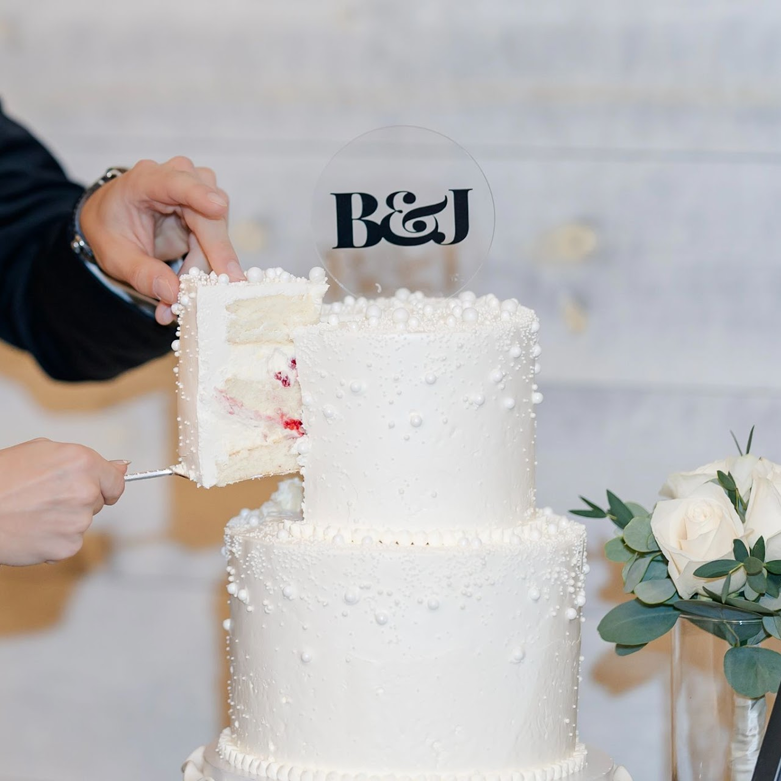 A person cutting a three-tier white wedding cake decorated with small white pearls and topped with a clear circular topper with the initials B&J.
