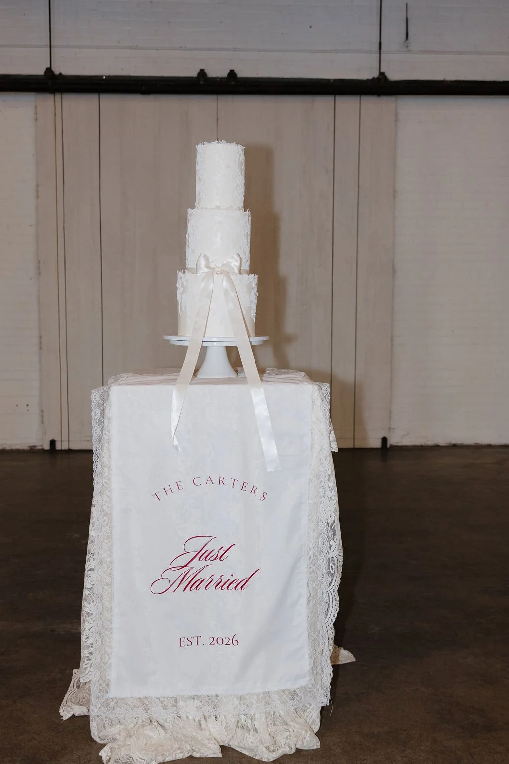 A white fabric sign that says "Just Married" on a hightop table with a lace tablecloth and a wedding cake on top.