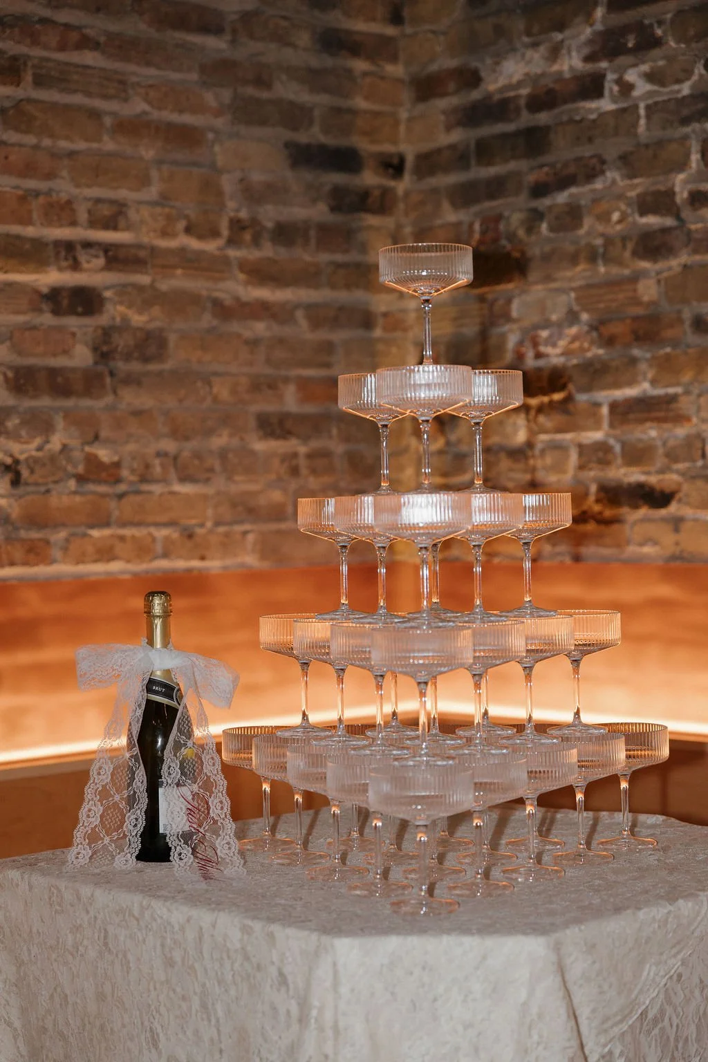 A champagne bottle with a lace ribbon bow on a table next to a champagne glass tower in a low lighting room with brick walls.