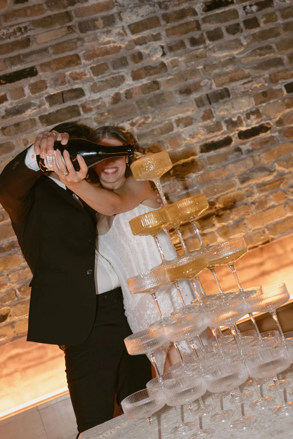 A bride and groom pouring champagne onto a champagne glass tower while smiling with a brick wall behind them.