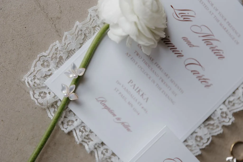 White wedding invitation with lace trim laid on a concrete floor with a white flower and earrings rested on top