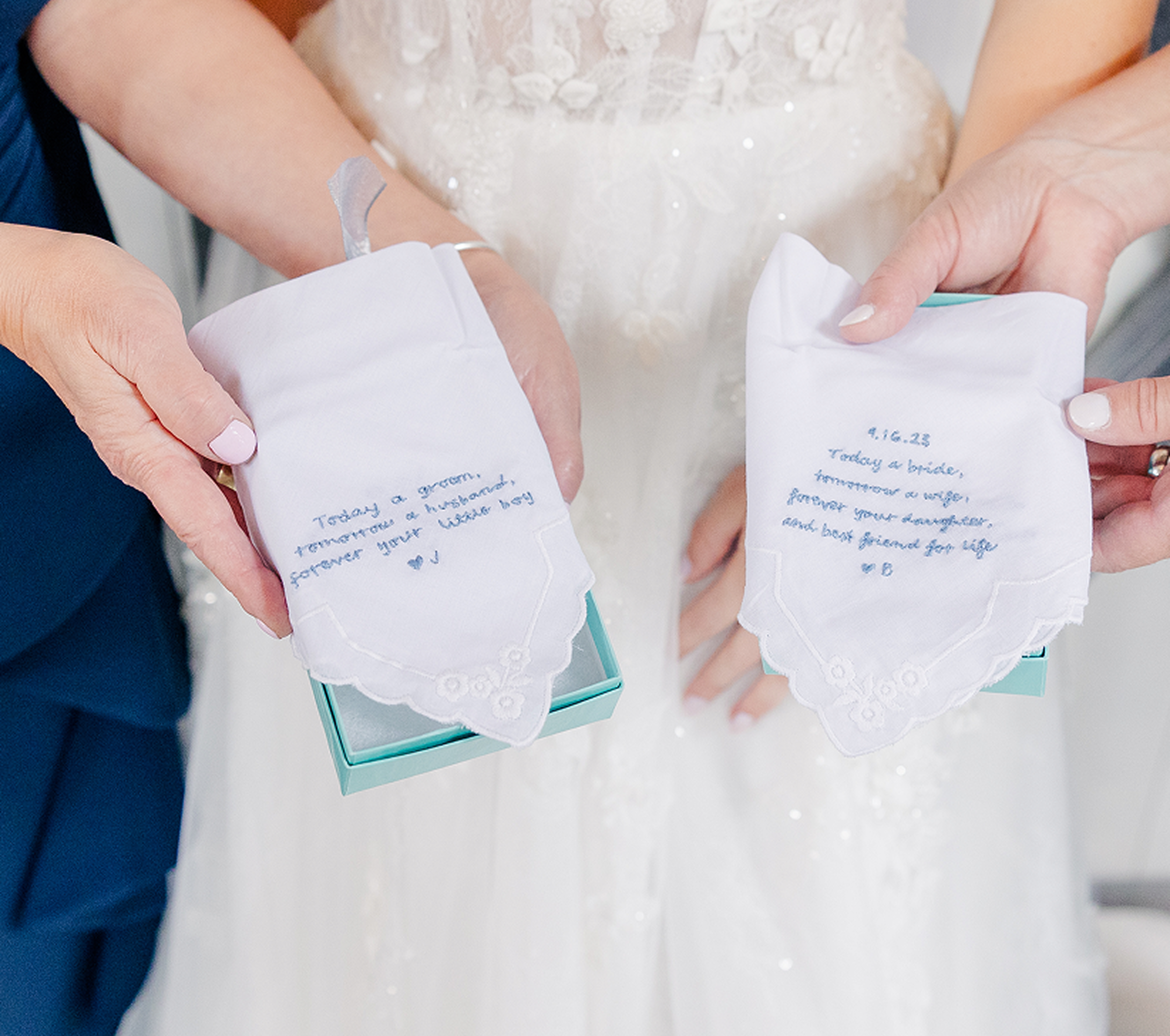 Two women holding embroidered handkerchiefs with blue thread spelling out notes from a bride and groom.