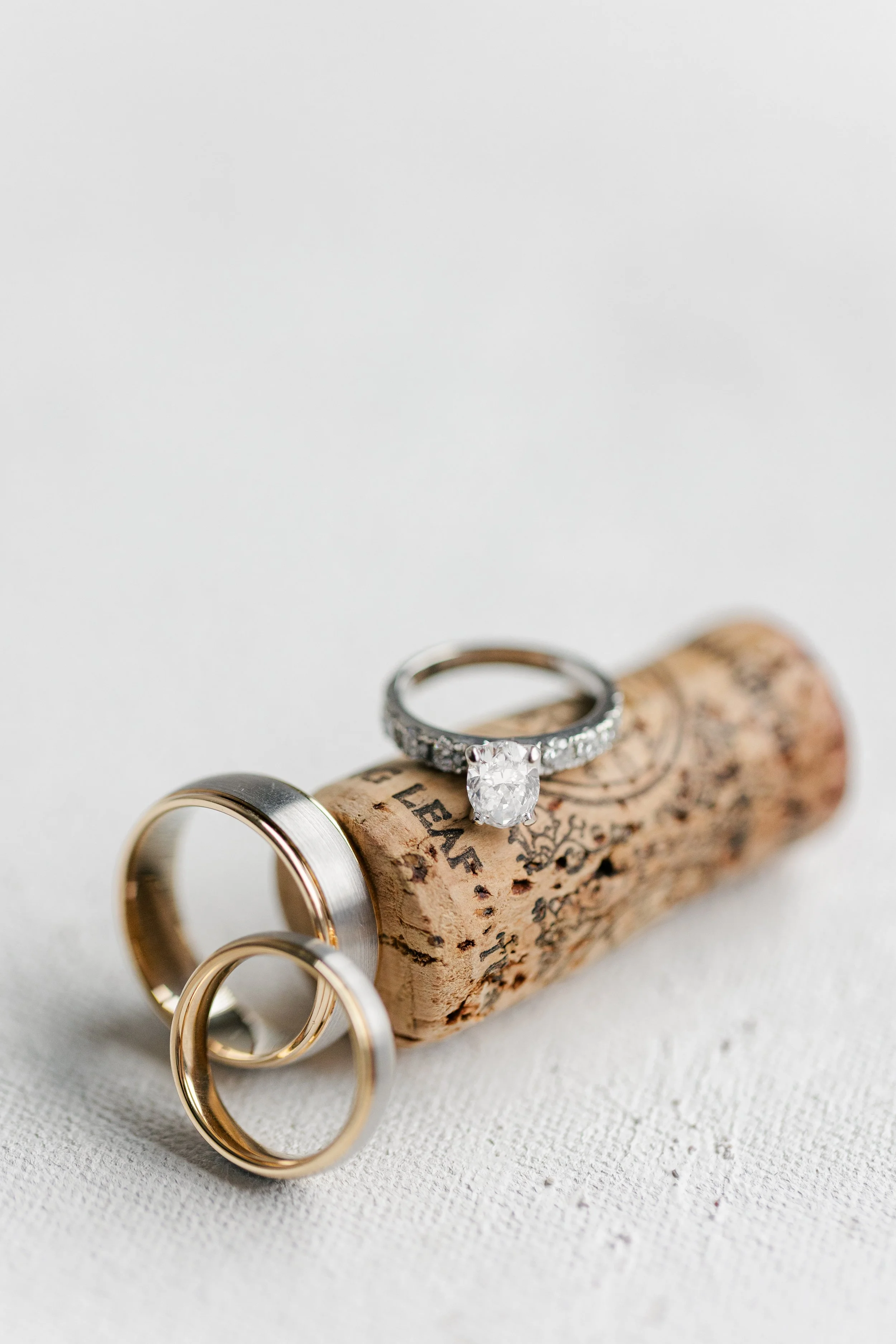 Wedding rings, including a diamond engagement ring and two gold bands, resting on a cork in front of a white fabric background.