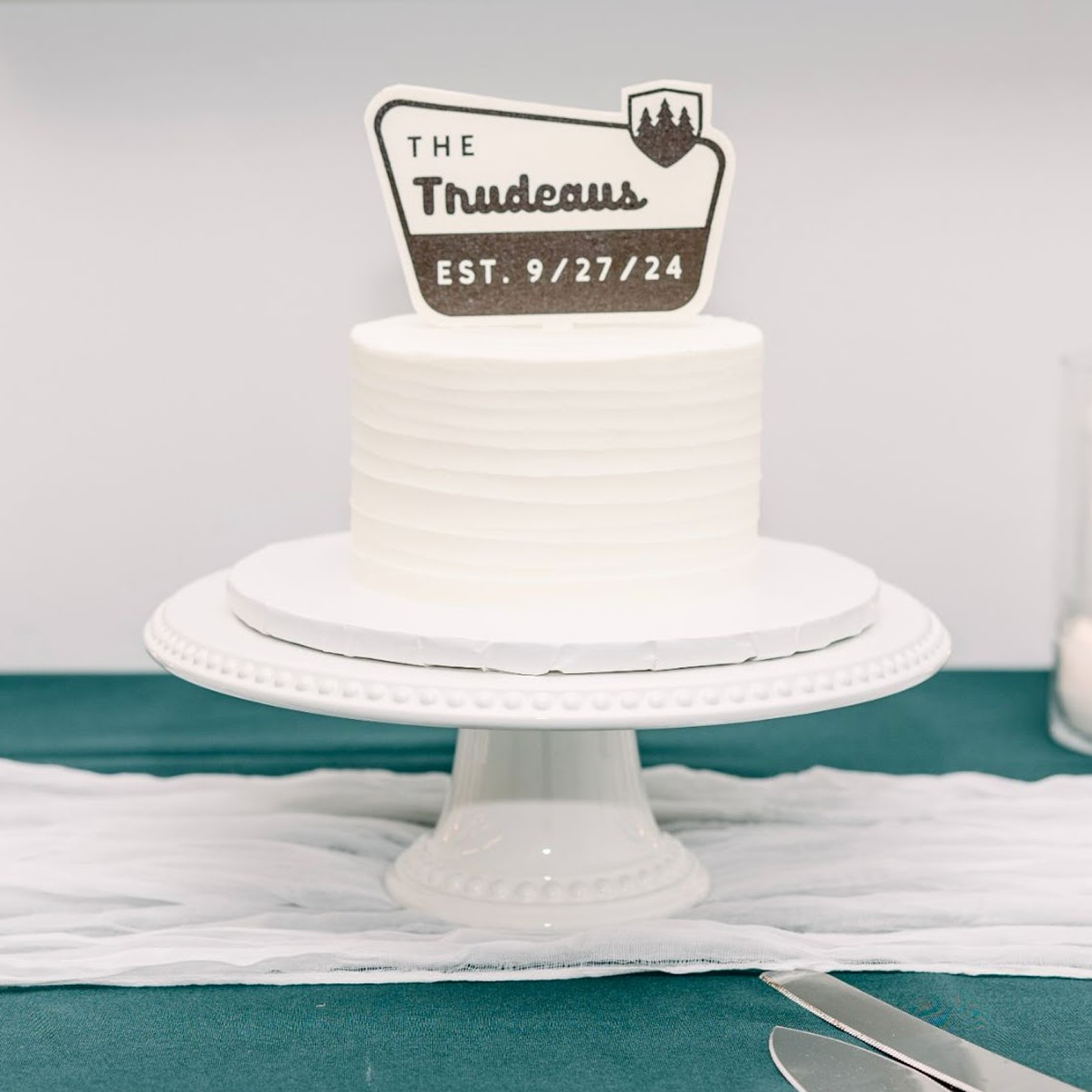 White wedding cake on a stand with a wooden cake topper on top in the style of a national parks welcome sign.