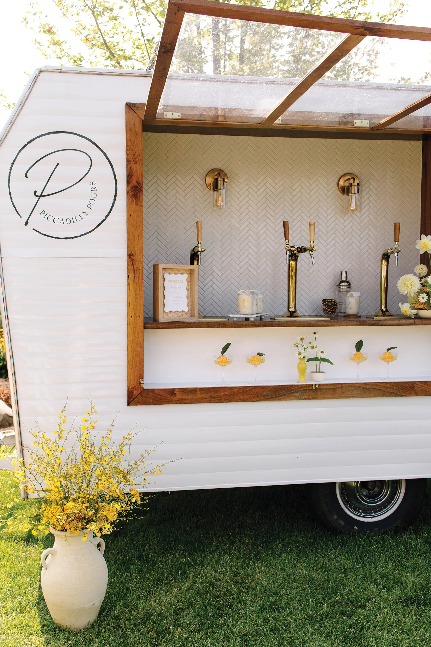 White bar camper with 3 taps, cocktails resting on the bar top, and a vase of yellow flowers in front