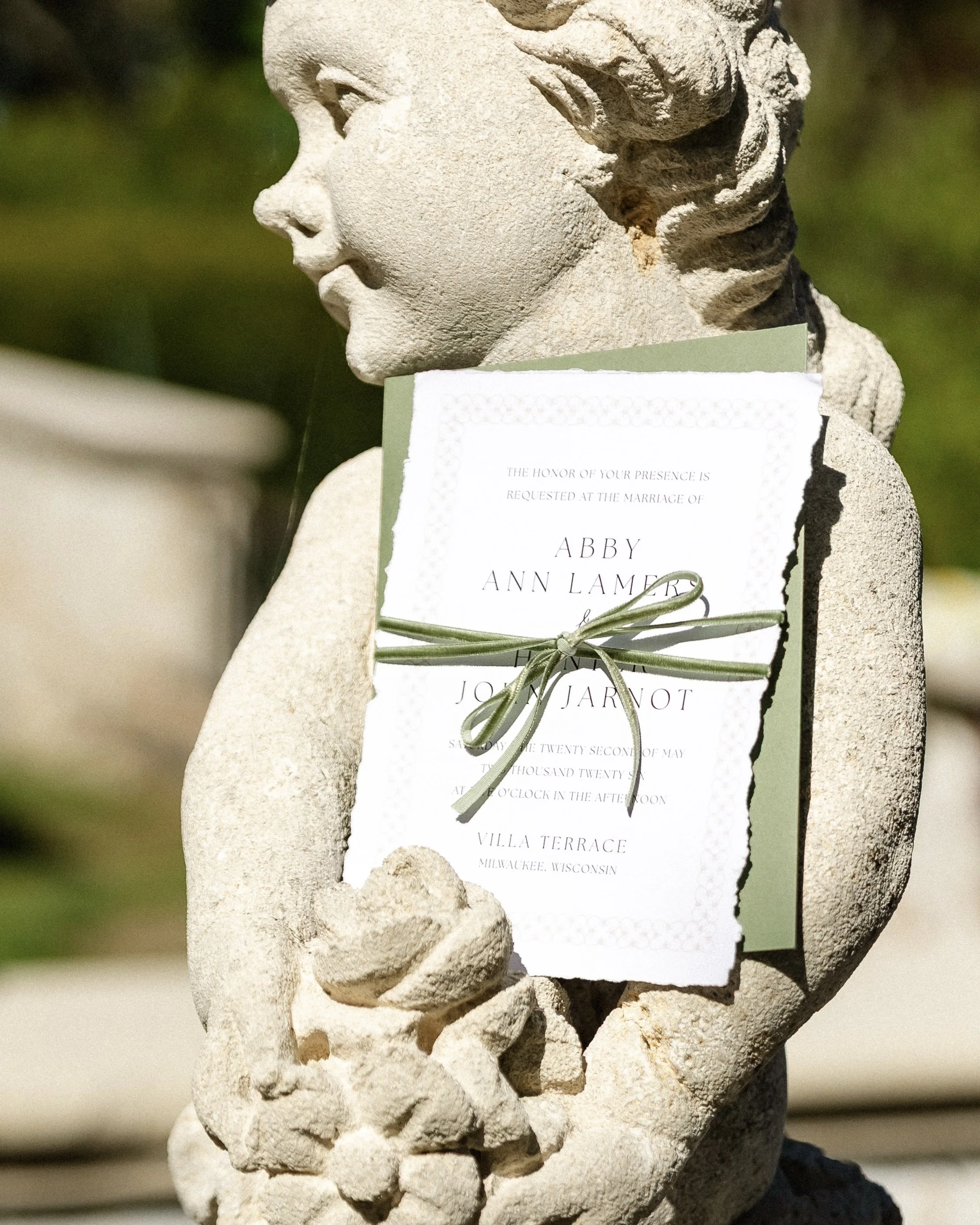 A ripped edge wedding invitation tied with sage green ribbon resting on a stone statue.