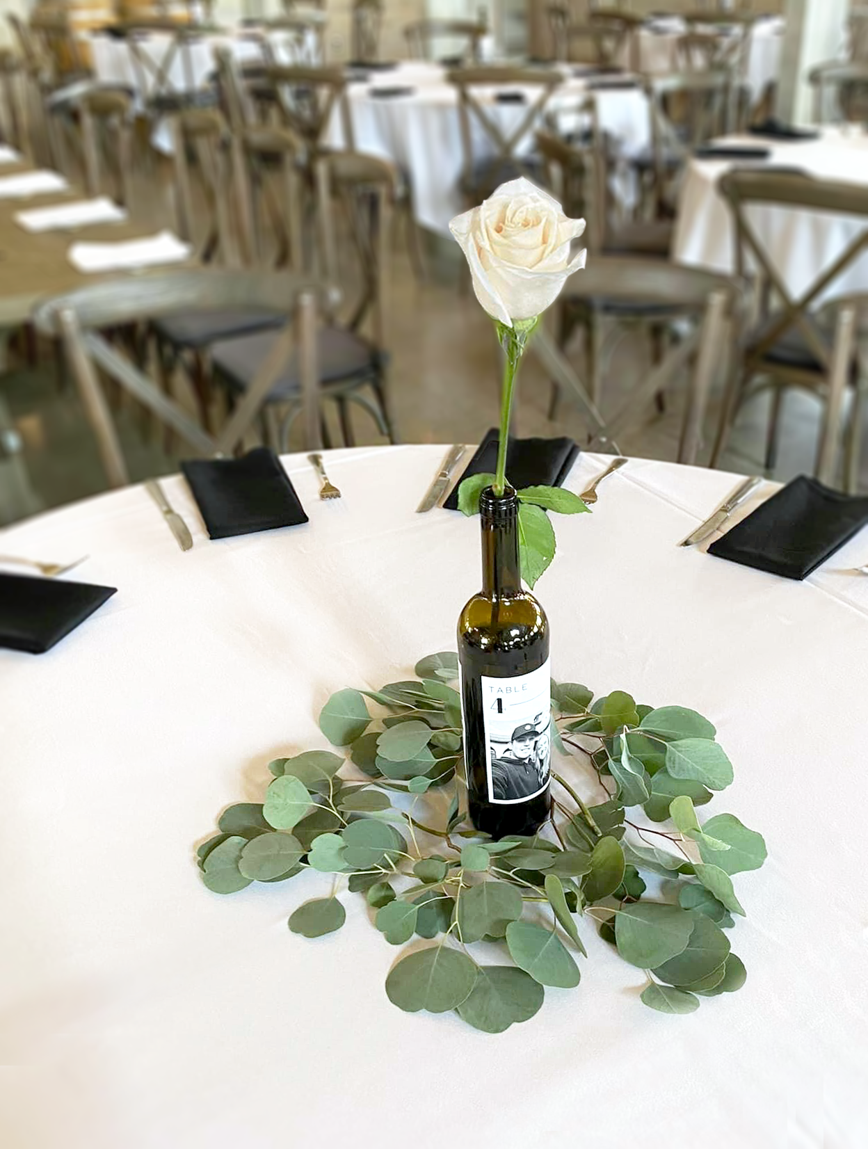 A wine bottle with a black and white photograph and the number 4 filled with a single white rose on a white tablecloth surrounded by eucalyptus branches.