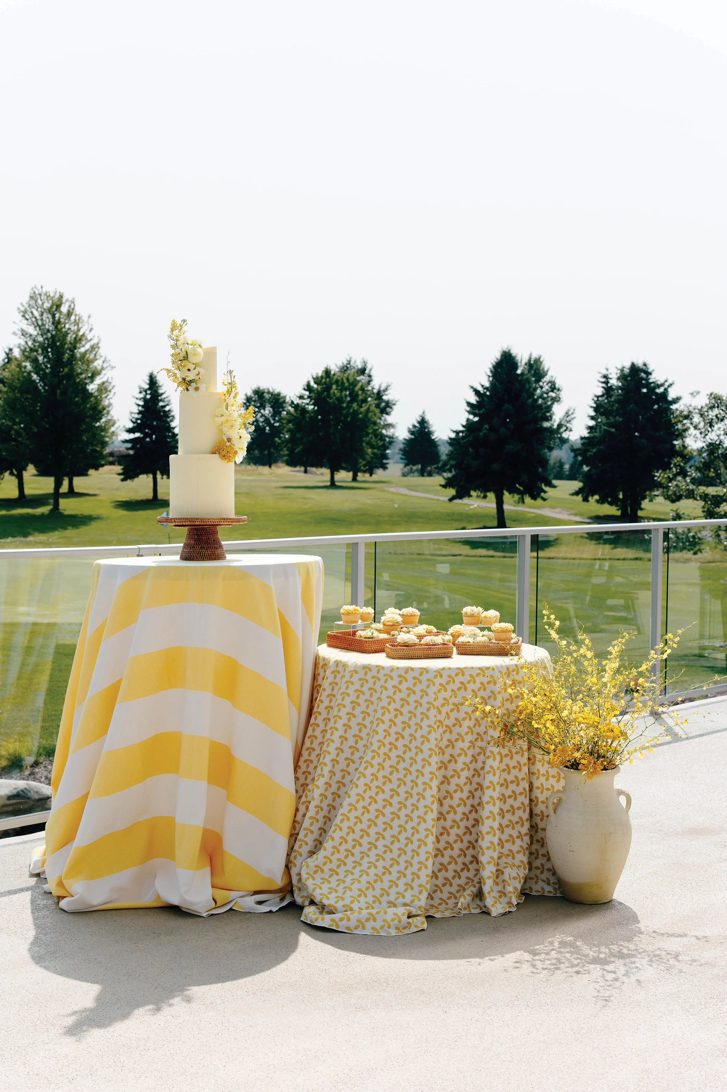 Two high top tables with different yellow printed tablecloths and a wedding cake and cupcakes on top