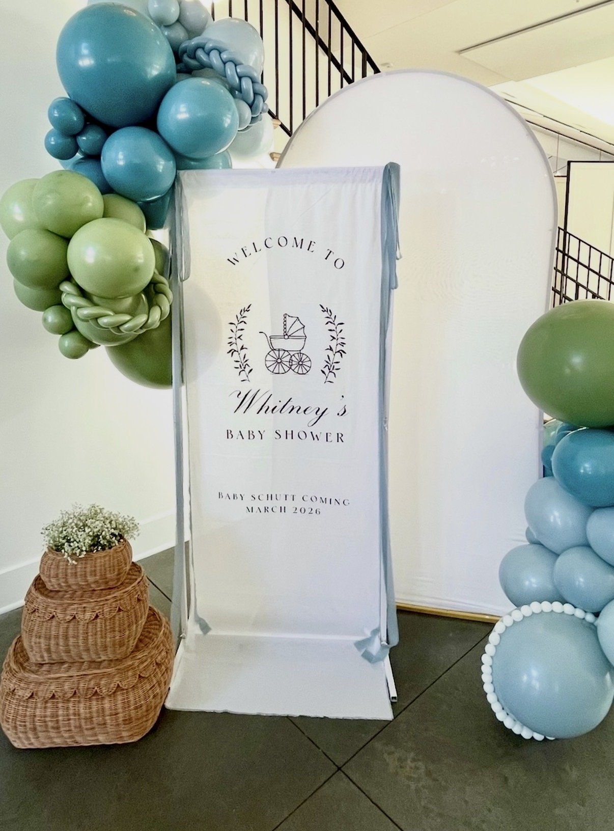 A white fabric welcome sign for a baby shower with blue ribbon surrounded by green and blue ribbons