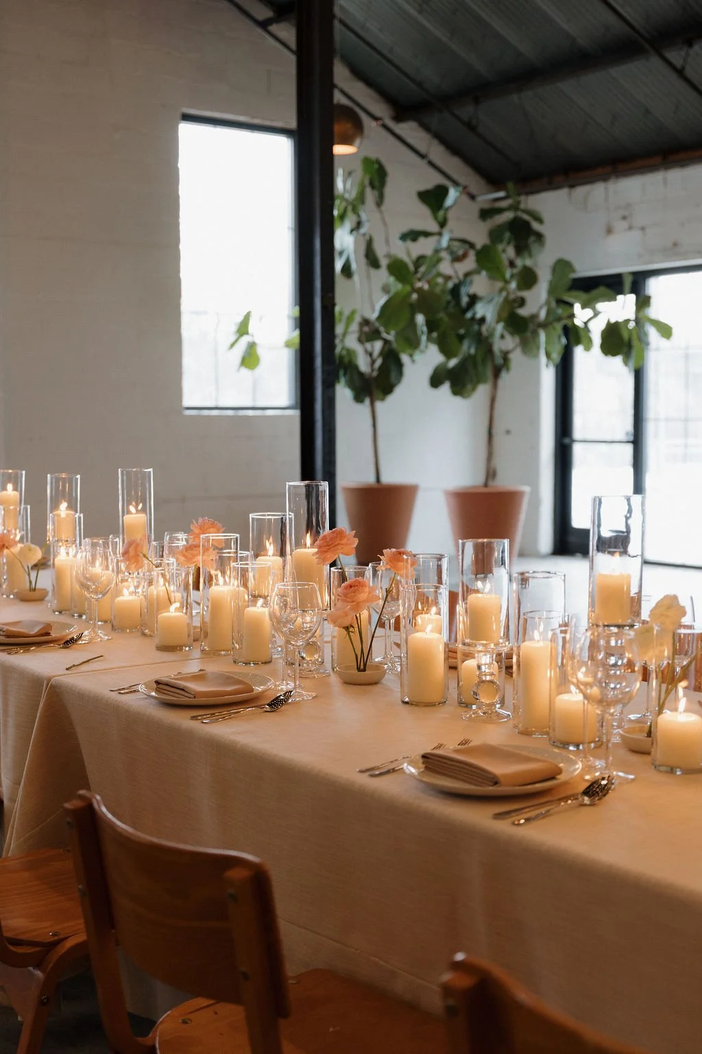 A wedding reception table with lit candles down the center and tall green plants in the background.