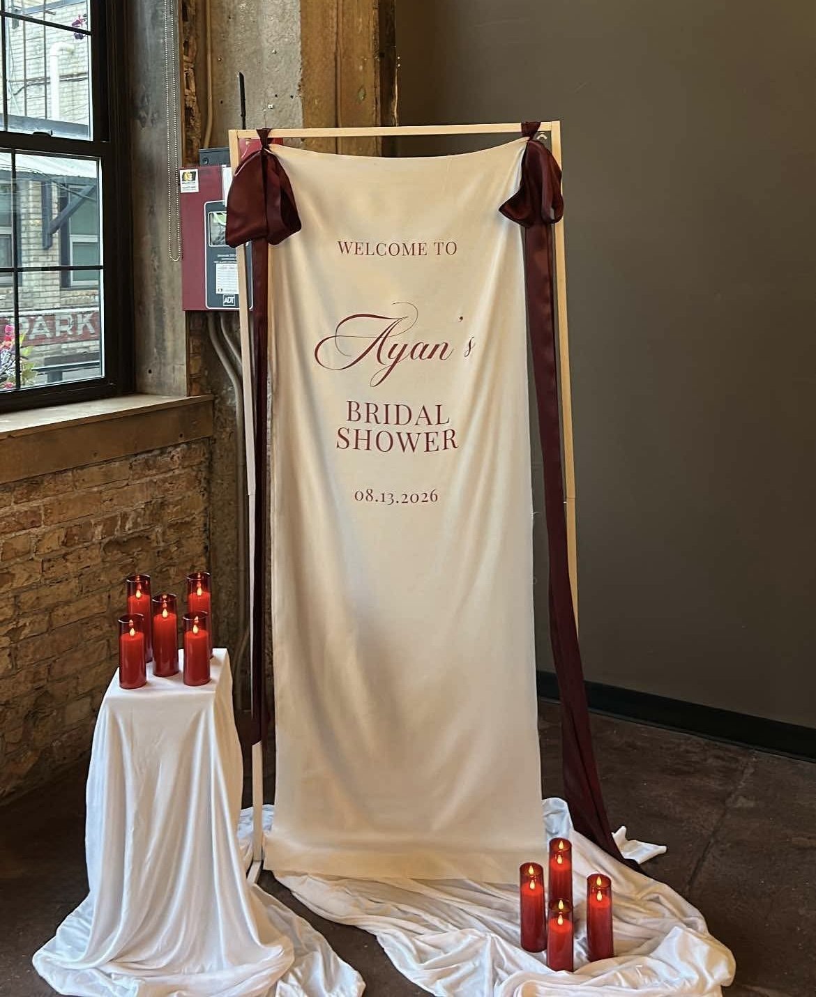 A white linen sign bridal shower sign tied onto a white stand with red ribbon and surrounded by red candles and draped white fabric.