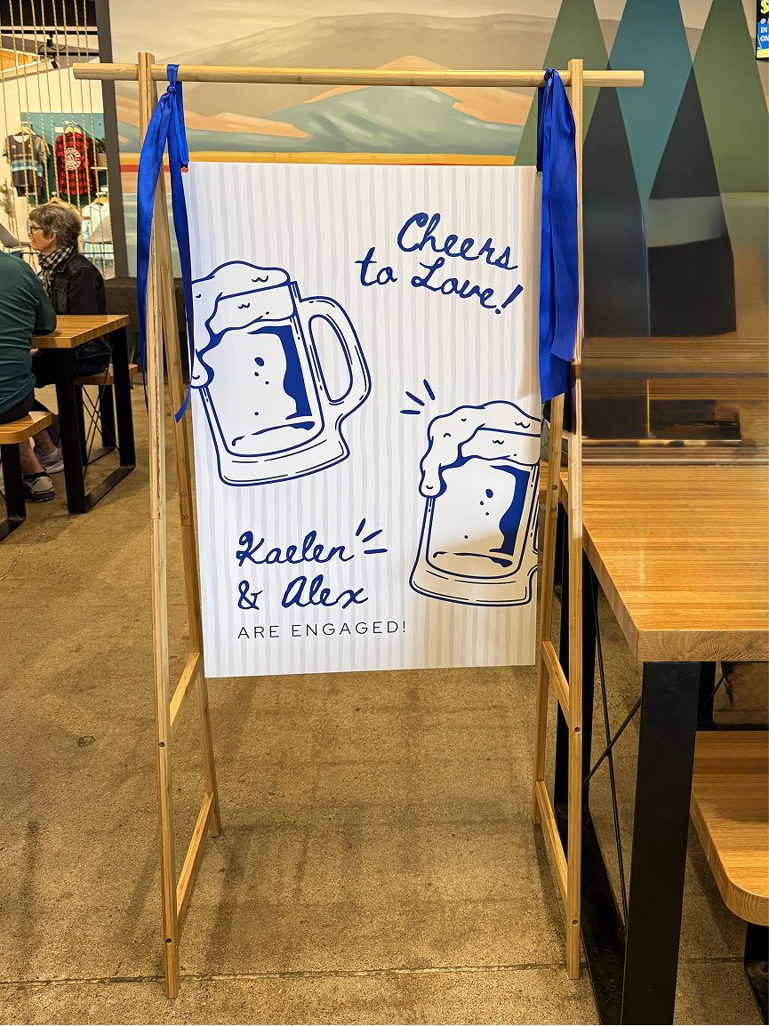 Blue and white striped sign that says "Cheers to Love" hanging from a wooden sign stand.