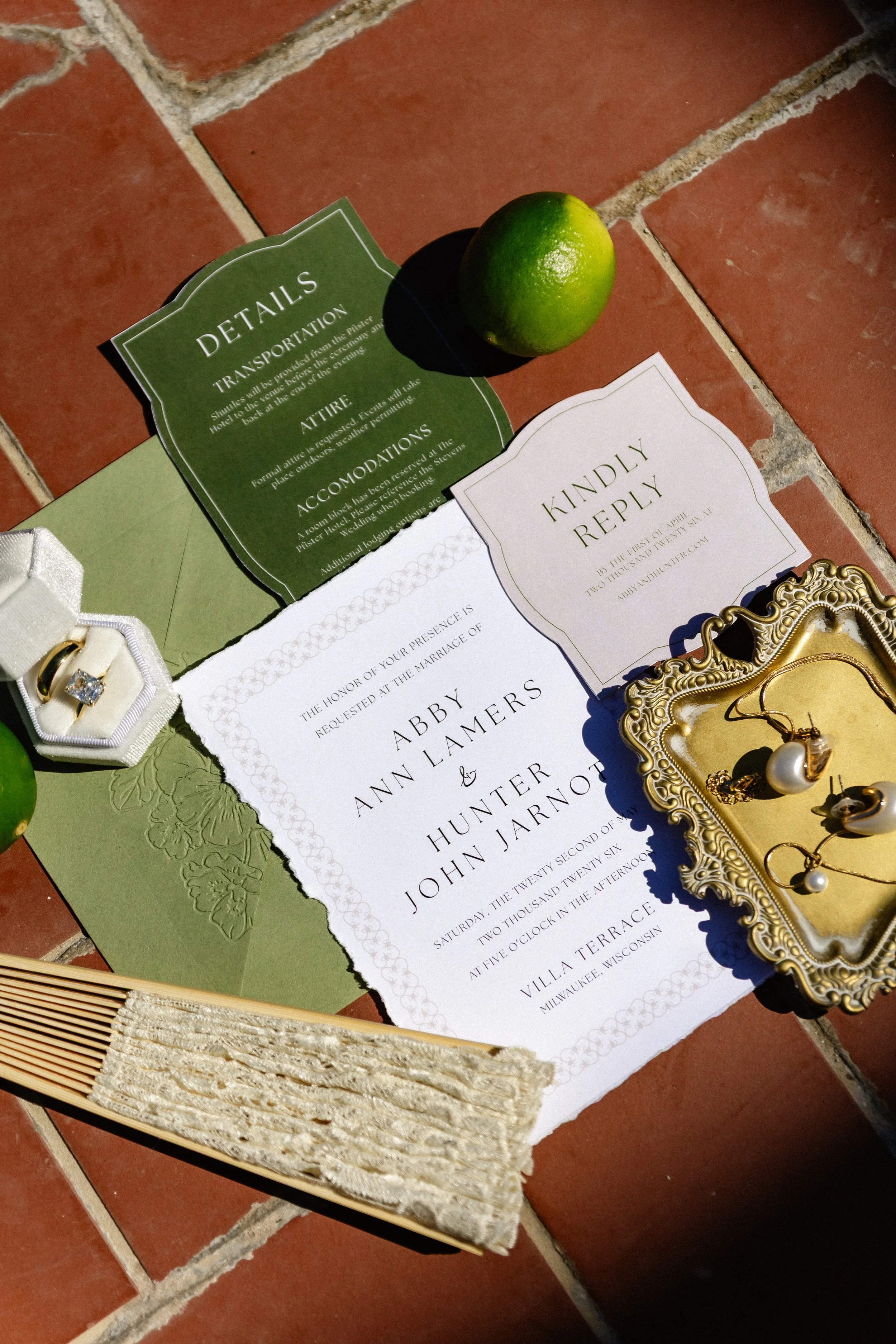 A white and green invitation suite flat lay resting on terracotta tiles. 