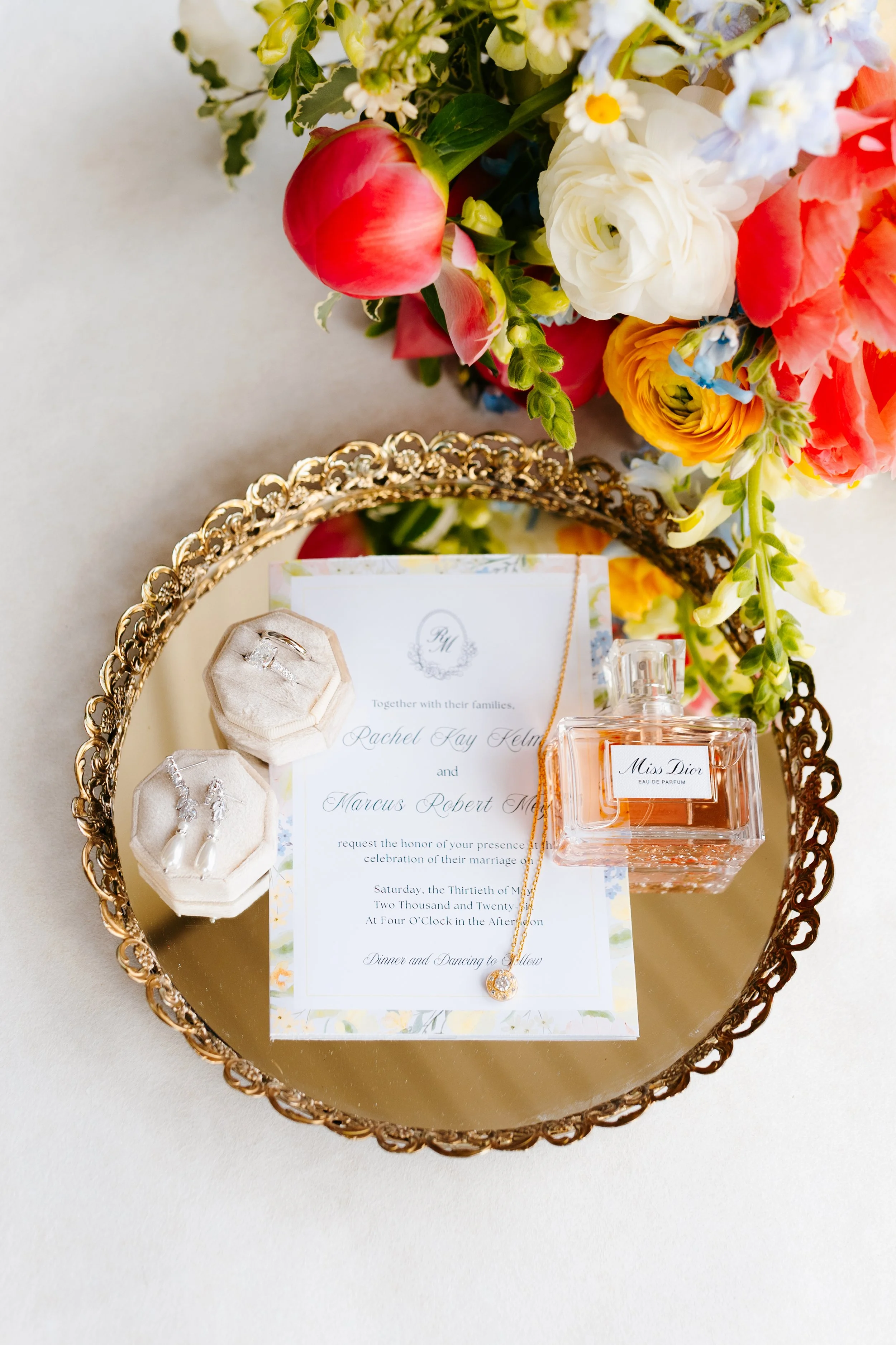 Wedding invitation with a floral border resting on a gold mirrored tray with a bottle of perfume, a ring box, and a floral bouquet nearby. 