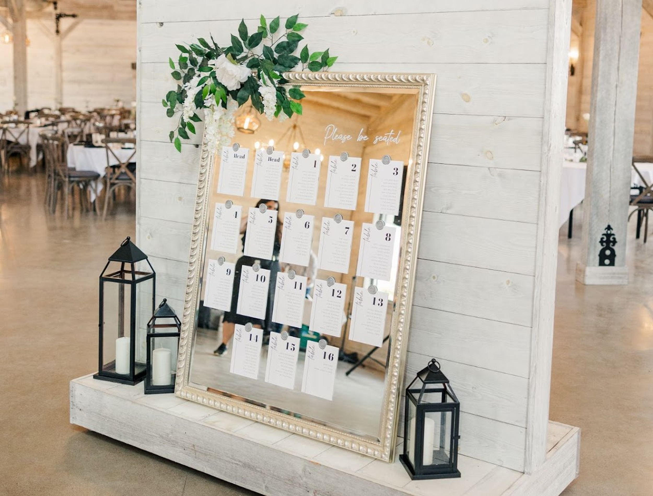 A large wedding seating chart mirror with paper cards attached with wax seals, all sitting on a large wooden stand with black lanterns decorating the base.