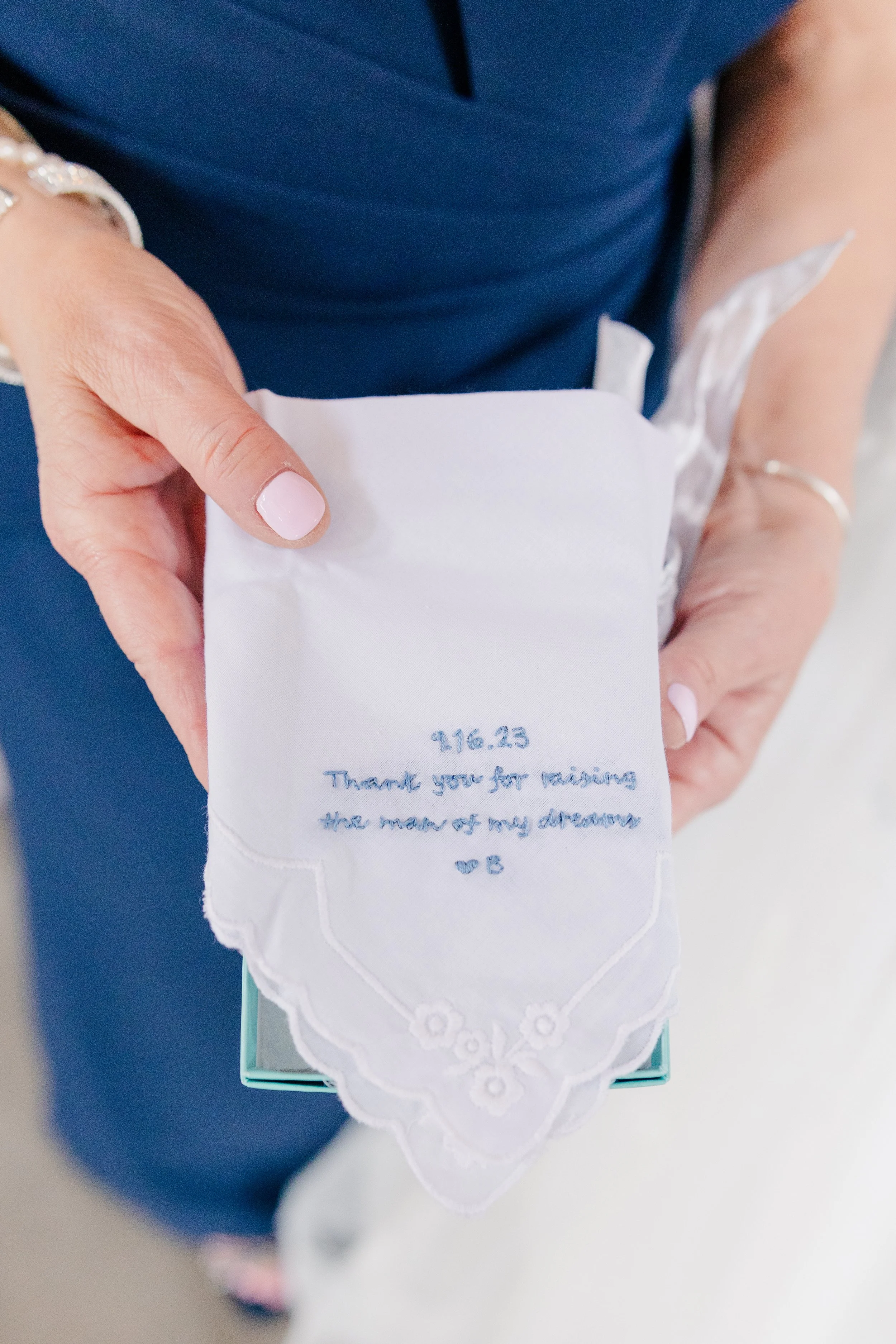 A woman holding a white handkerchief with embroidered text that reads '9.16.23 Thank you for raising the man of my dreams ♥ B'.