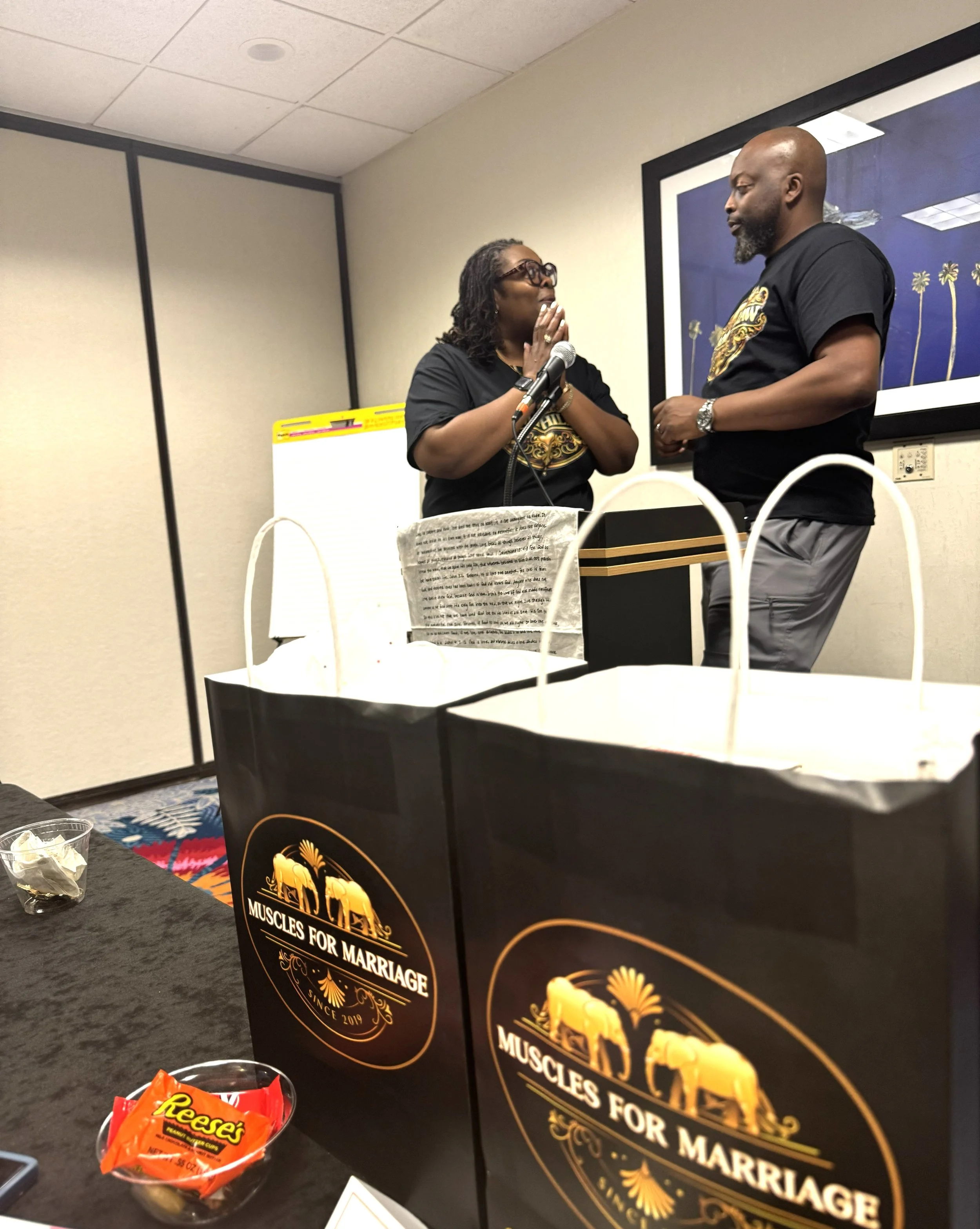 Two people, a man and a woman, are smiling and talking in a conference room. They are wearing matching black T-shirts, and the man is gesturing with his hands while the woman looks at him. There is a large TV screen behind them, and the room has blue curtains and a carpet with a geometric pattern.