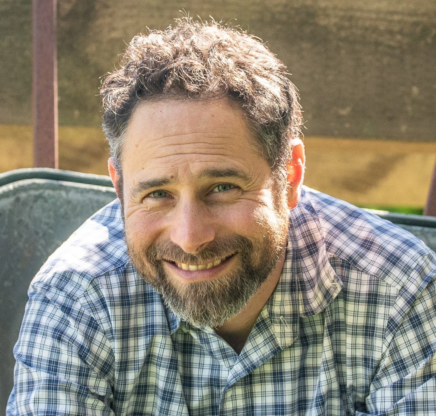 A man with curly brown hair and a beard smiling outdoors, wearing a plaid shirt.