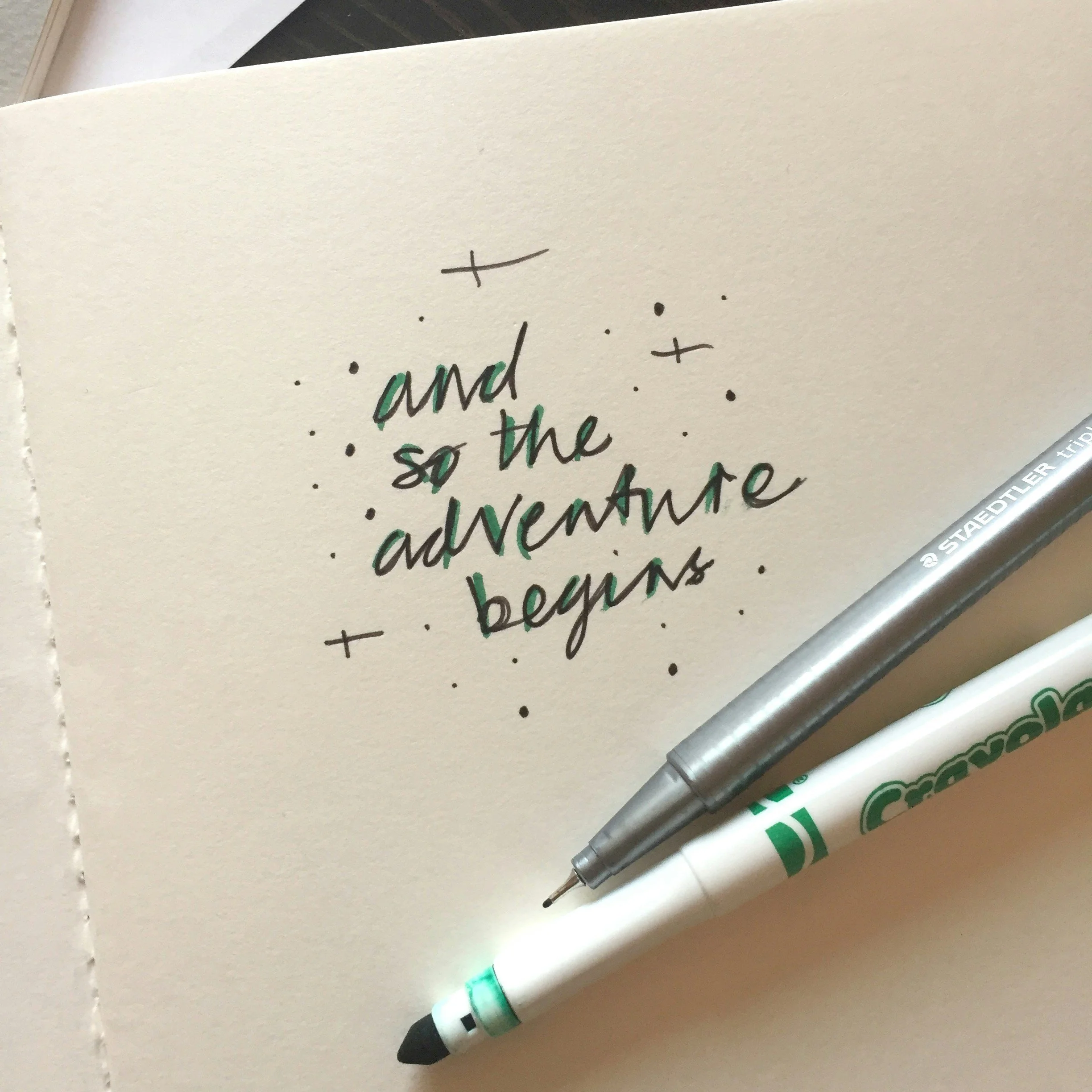 A notebook page with handwritten text saying 'and so the adventure begins' surrounded by small decorative dots and plus signs, with a lab pen displaying a green LED light and a white marker with a green logo lying on top.