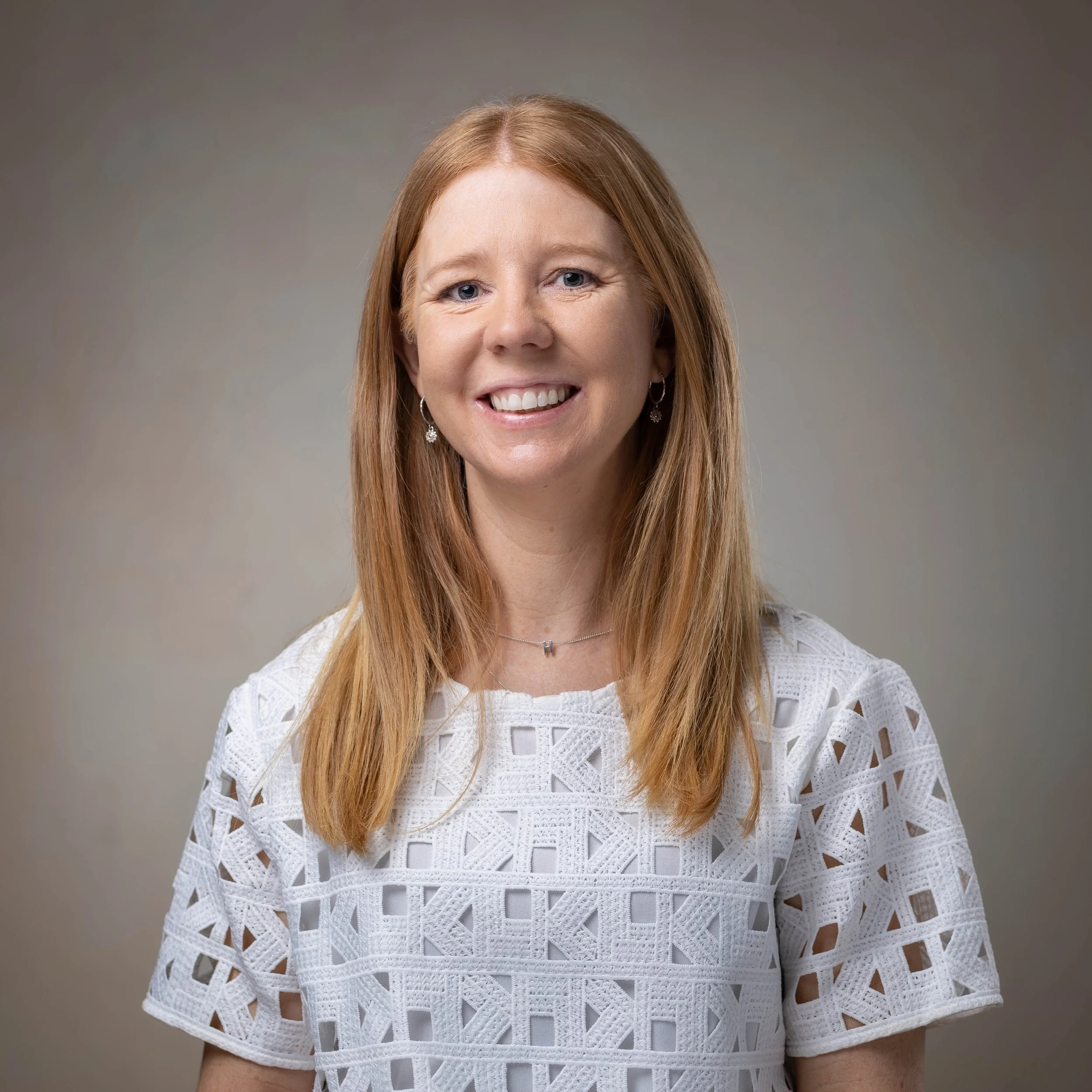 A smiling woman with long red hair, wearing a white lace-patterned top, earrings, and a necklace, against a neutral gray background.