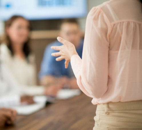 A person gesturing with their hands while speaking during a meeting, with others sitting at a table in the background.
