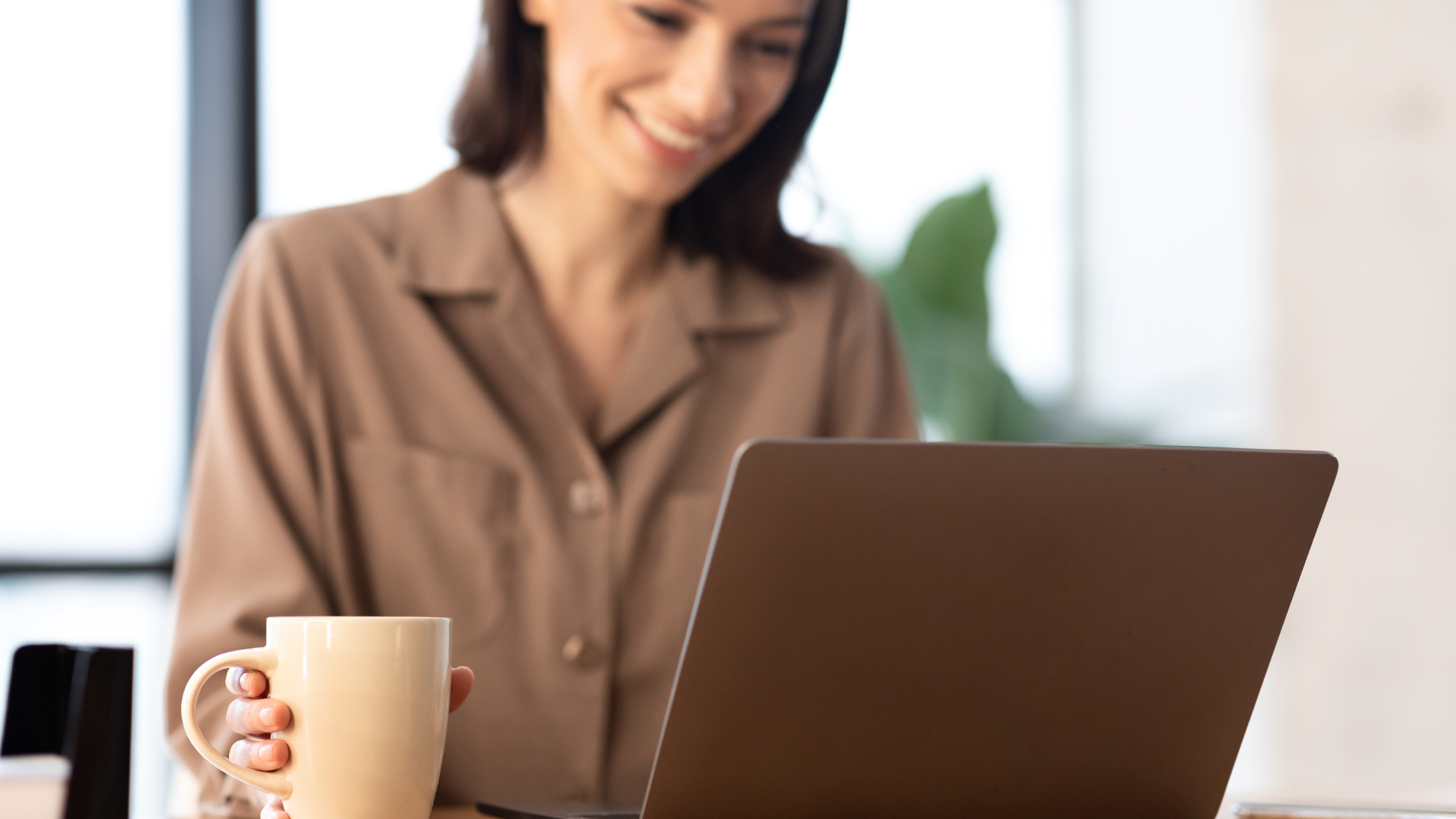 A woman in a tan blouse smiling while looking at her laptop, holding a white mug in her left hand, with a blurred background including a large window and greenery.