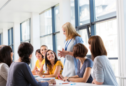 A group of women in a business meeting, sitting at a table together, with a woman standing and speaking to them, inside a bright conference room with large windows.