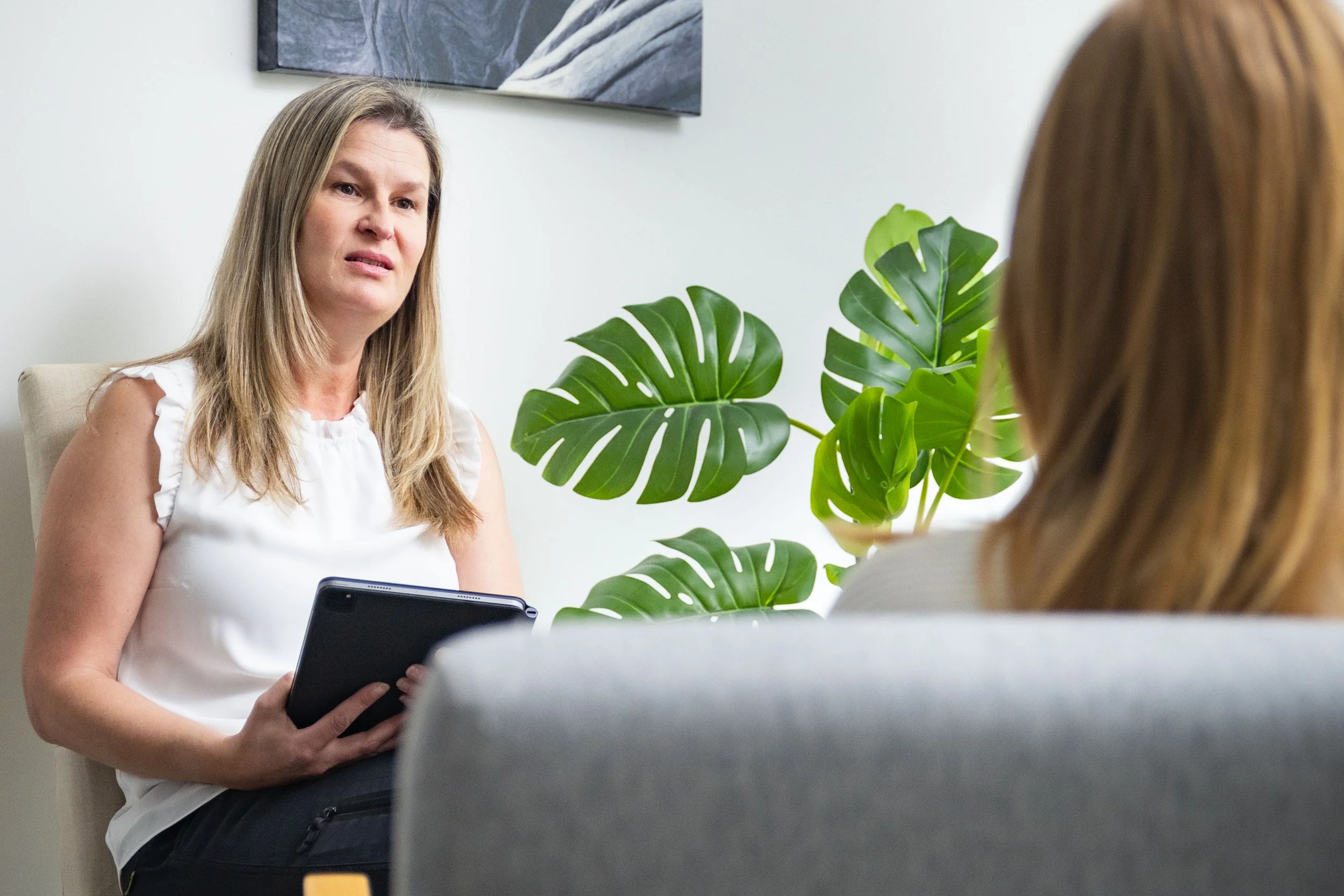 A woman with long blonde hair in a white sleeveless top sitting on a beige chair during a conversation with another woman with light brown hair whose back is turned. There's a large green monstera plant and a framed black and white photograph on the wall.