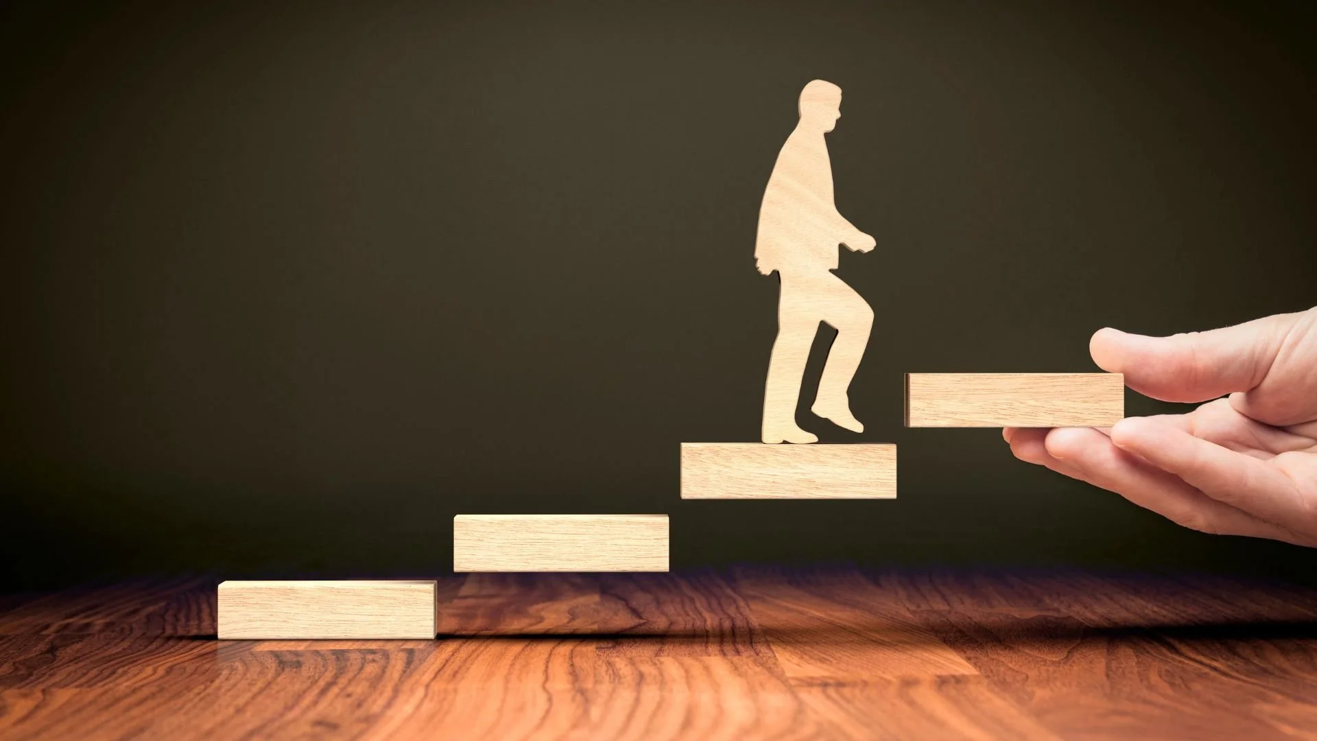 A hand placing a wooden block to complete a staircase of blocks, with a silhouette of a person walking up the stairs, representing growth or progress.