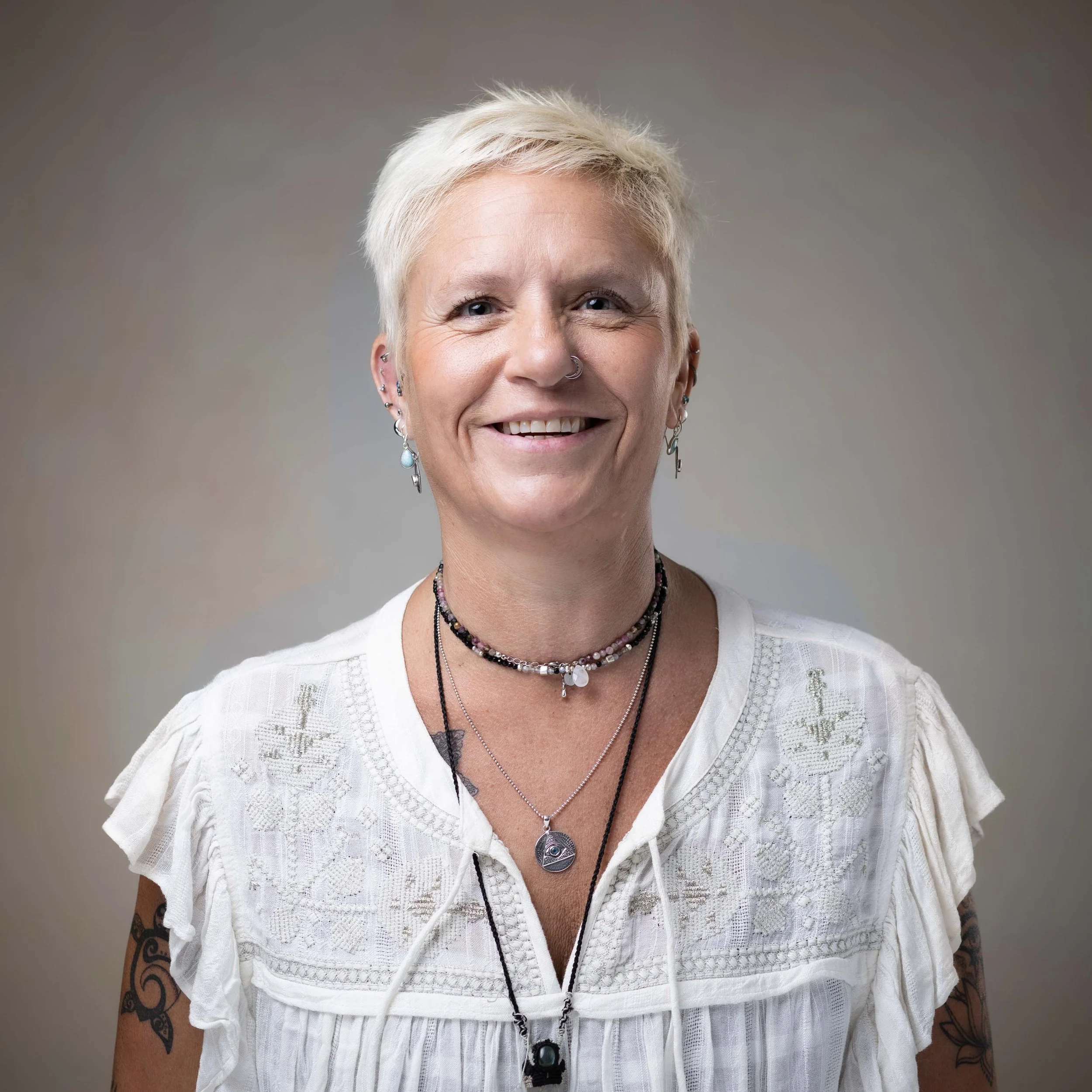 Portrait of a woman with short white hair, multiple earrings, and layered necklaces, smiling in front of a neutral background.