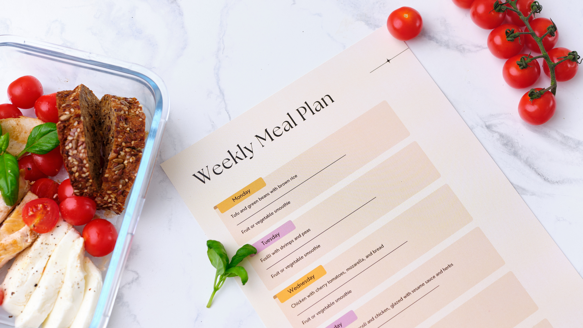 A weekly meal plan worksheet on a white surface, with cherry tomatoes, a basil leaf, and a glass container filled with sliced bread, cherry tomatoes, and mozzarella cheese nearby.