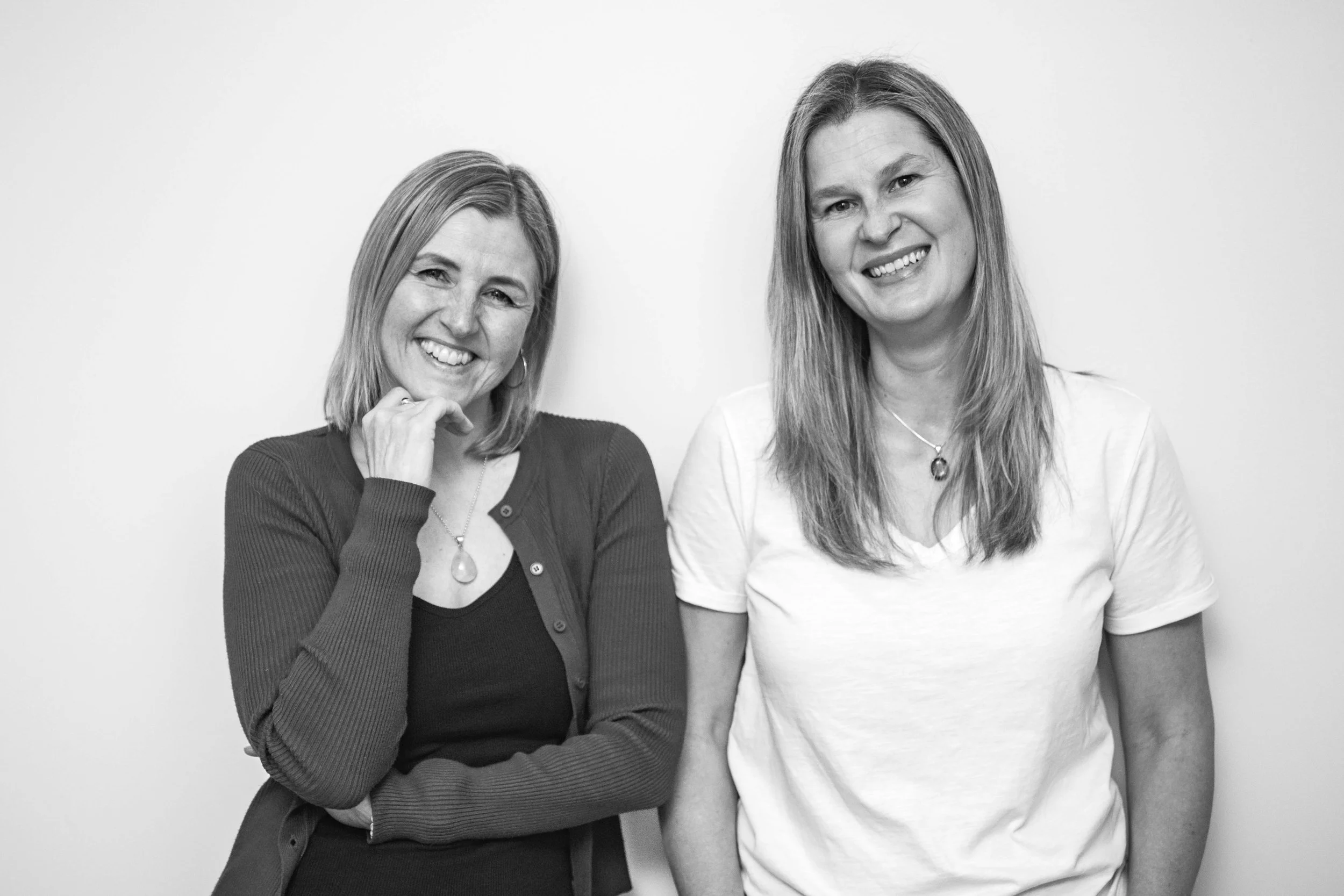 Two women standing side by side against a plain wall, smiling at the camera.