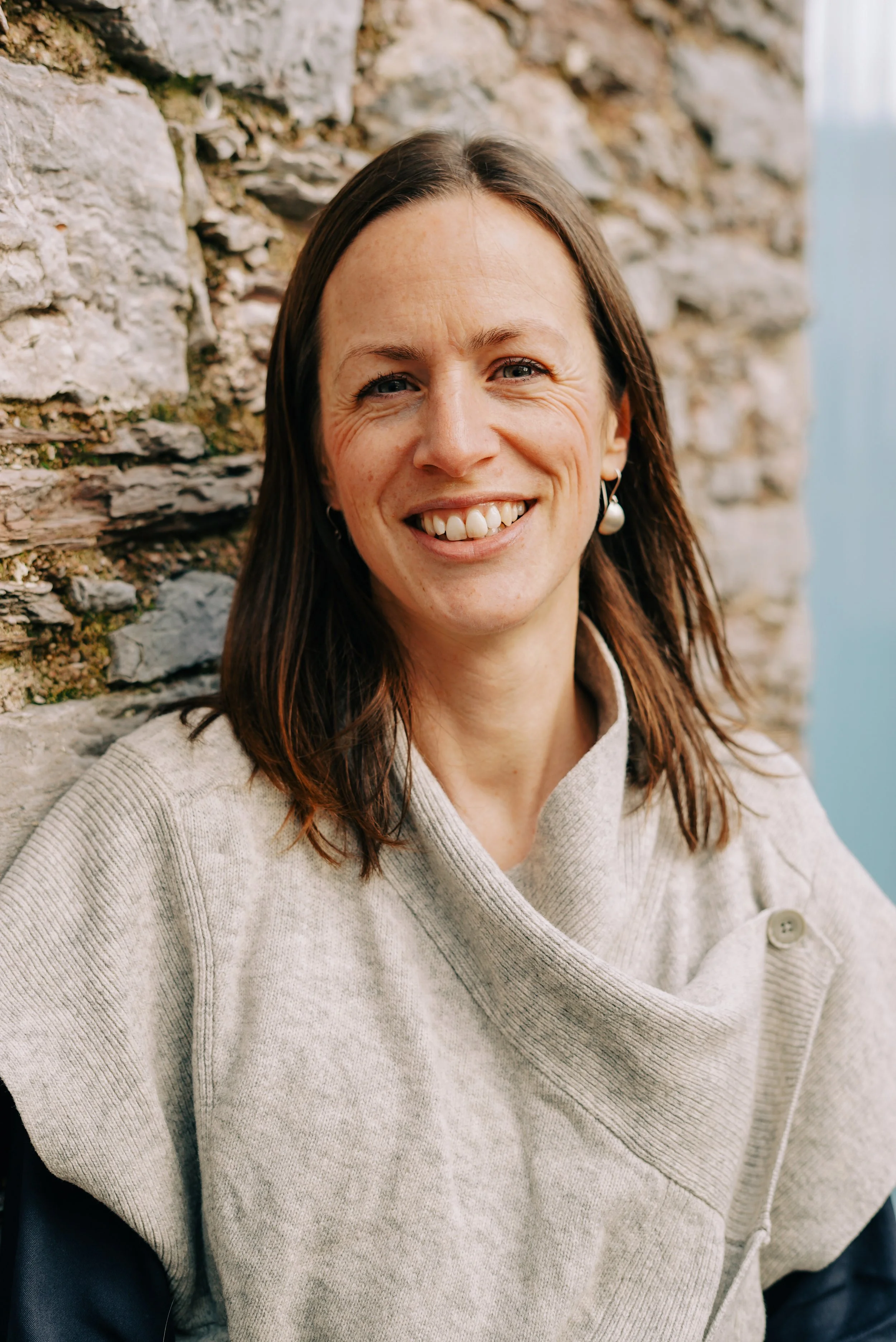 A woman with brown hair, smiling and showing teeth, standing against a stone wall outdoors.