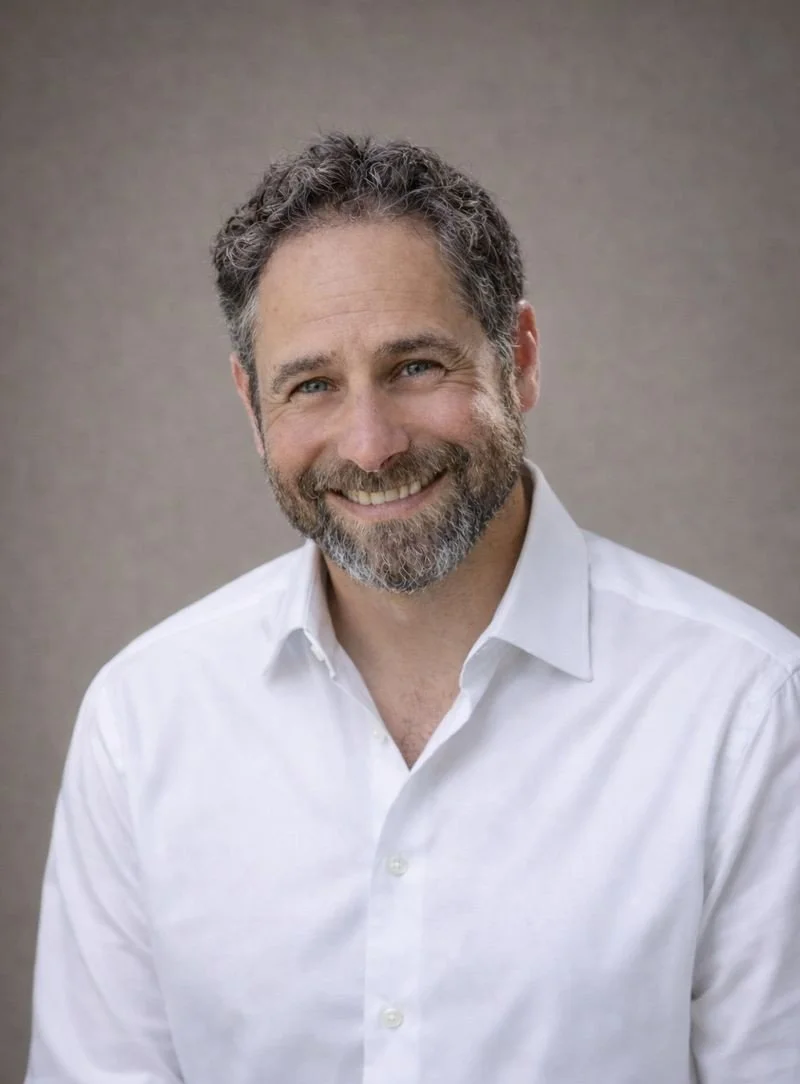 A smiling man with curly, salt-and-pepper hair and a beard, wearing a white dress shirt against a neutral background.