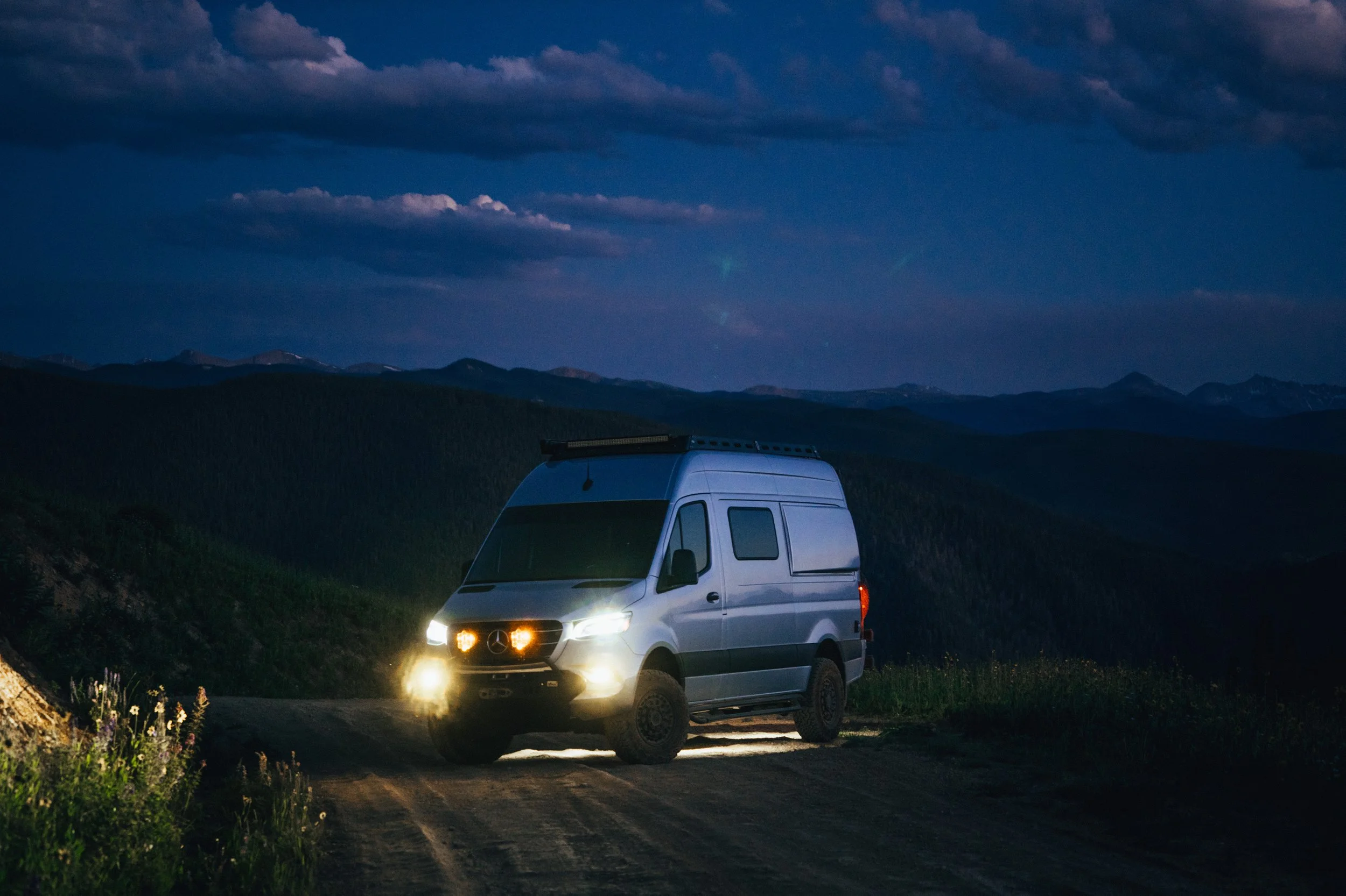 A silver van with its headlights on parked on a dirt road at night, with mountains and a cloudy sky in the background.