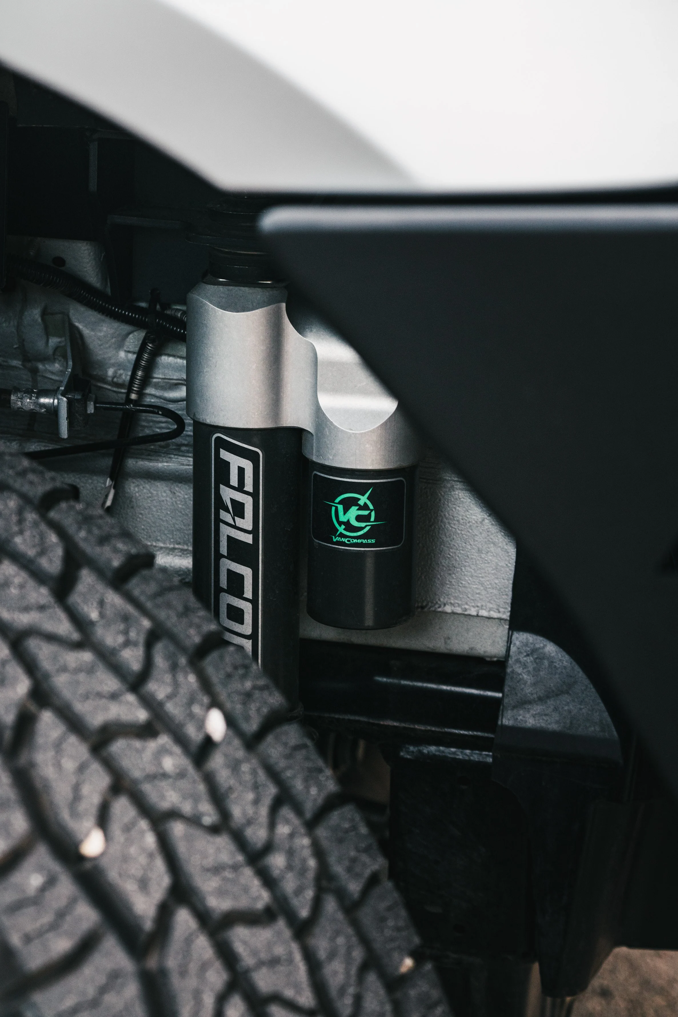 Close-up of a vehicle's suspension system, showing a coilover shock absorber with Felco branding and a VanCompass sticker, near a tire tread.