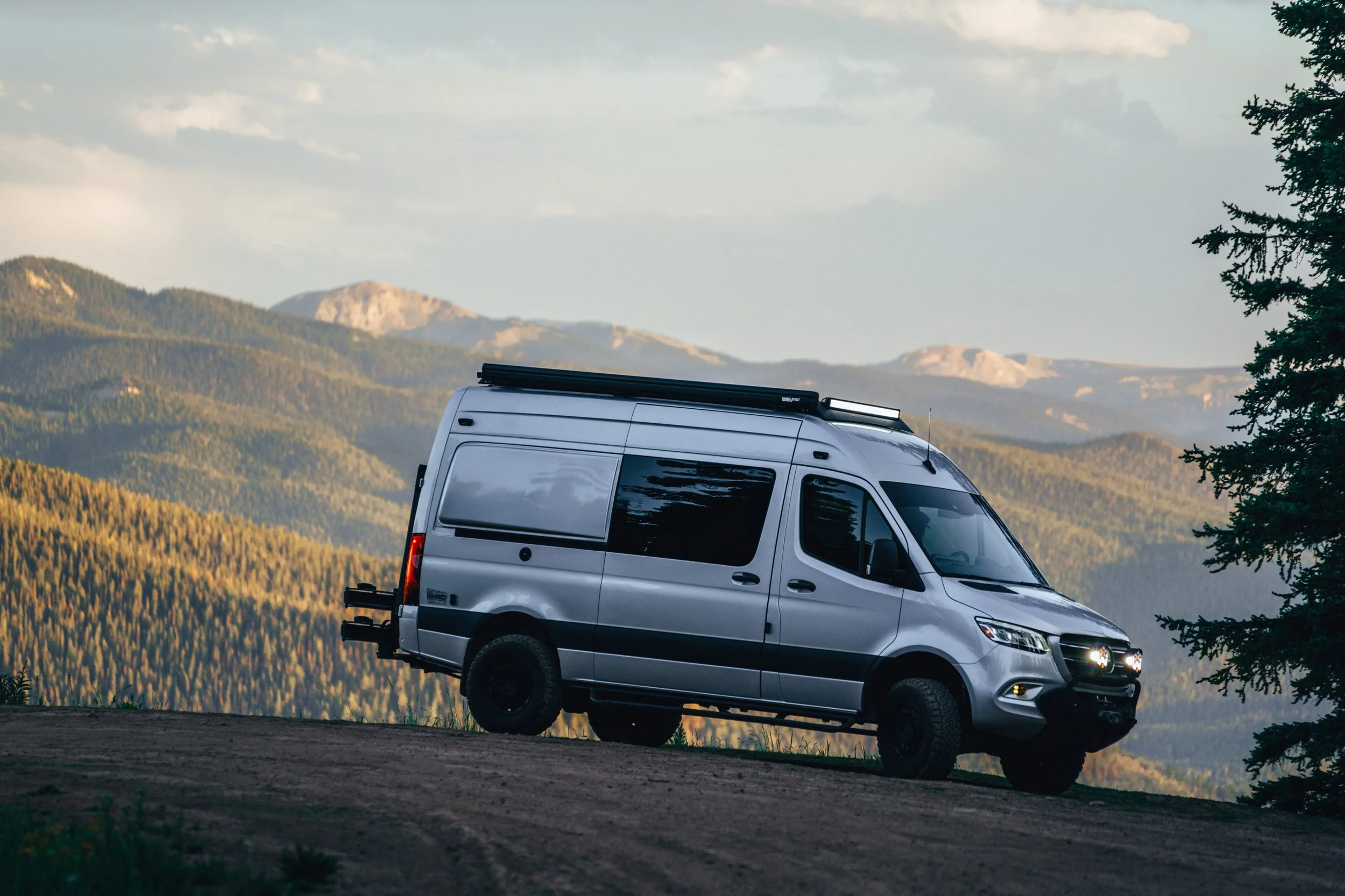Silver van parked on a dirt road in front of mountains and forest with a cloudy sky.