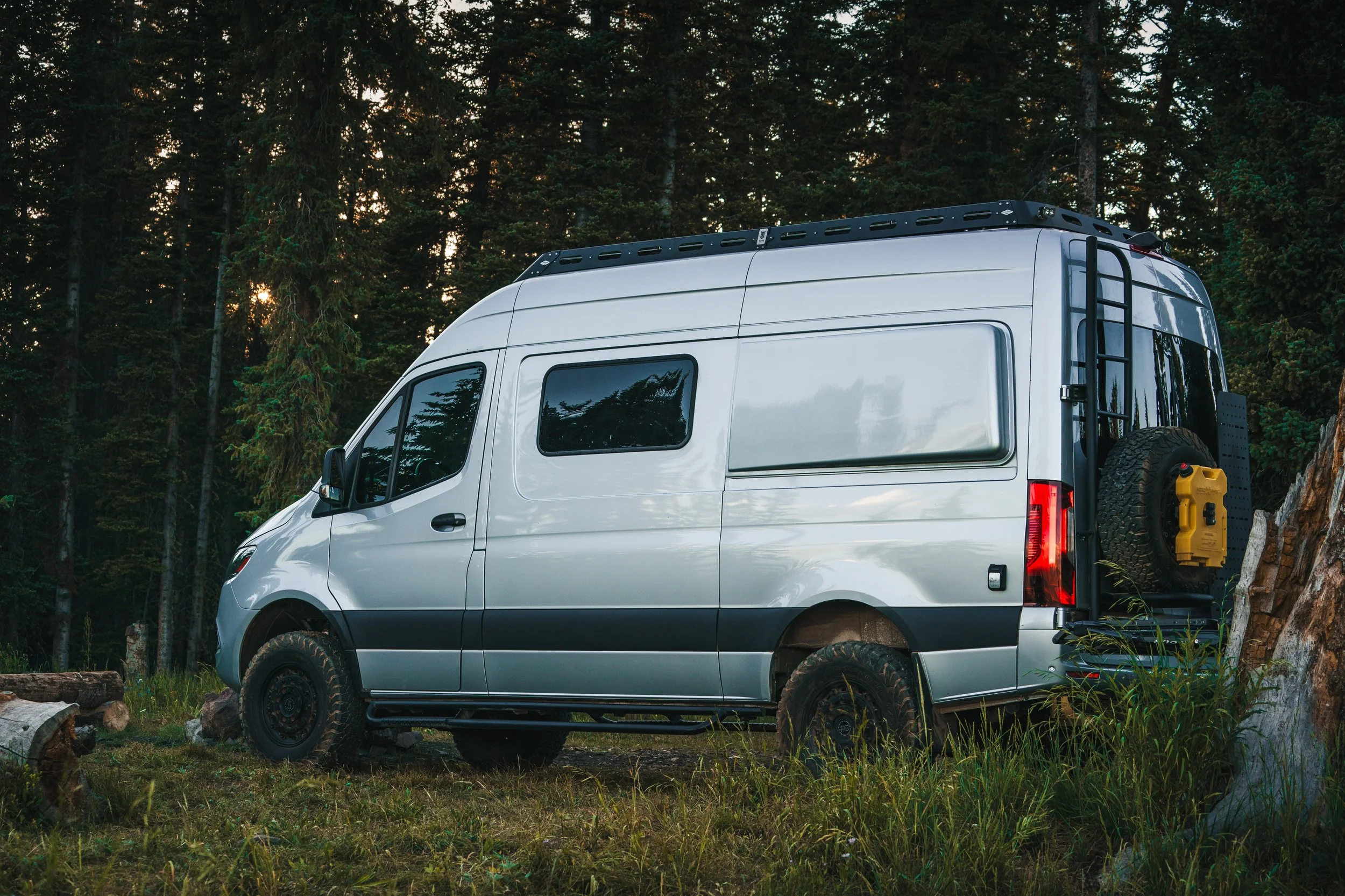 A silver camper van parked in a forested area with trees and grass, and a yellow container attached to the back of the vehicle.
