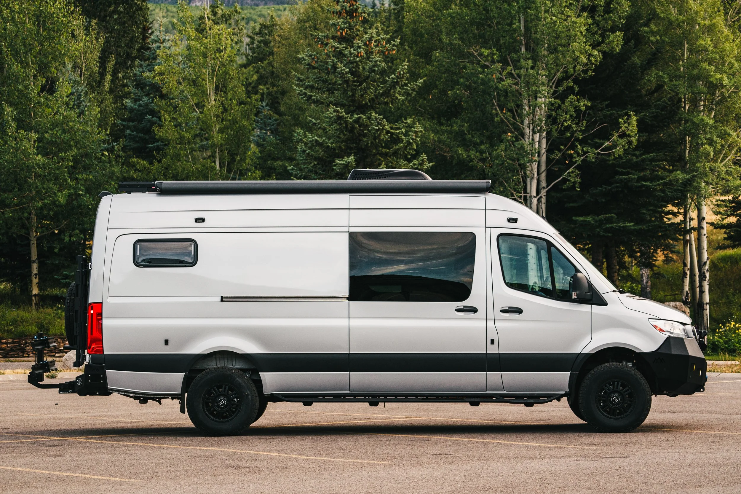 A silver Sprinter camper van parked on an empty parking lot surrounded by green trees.