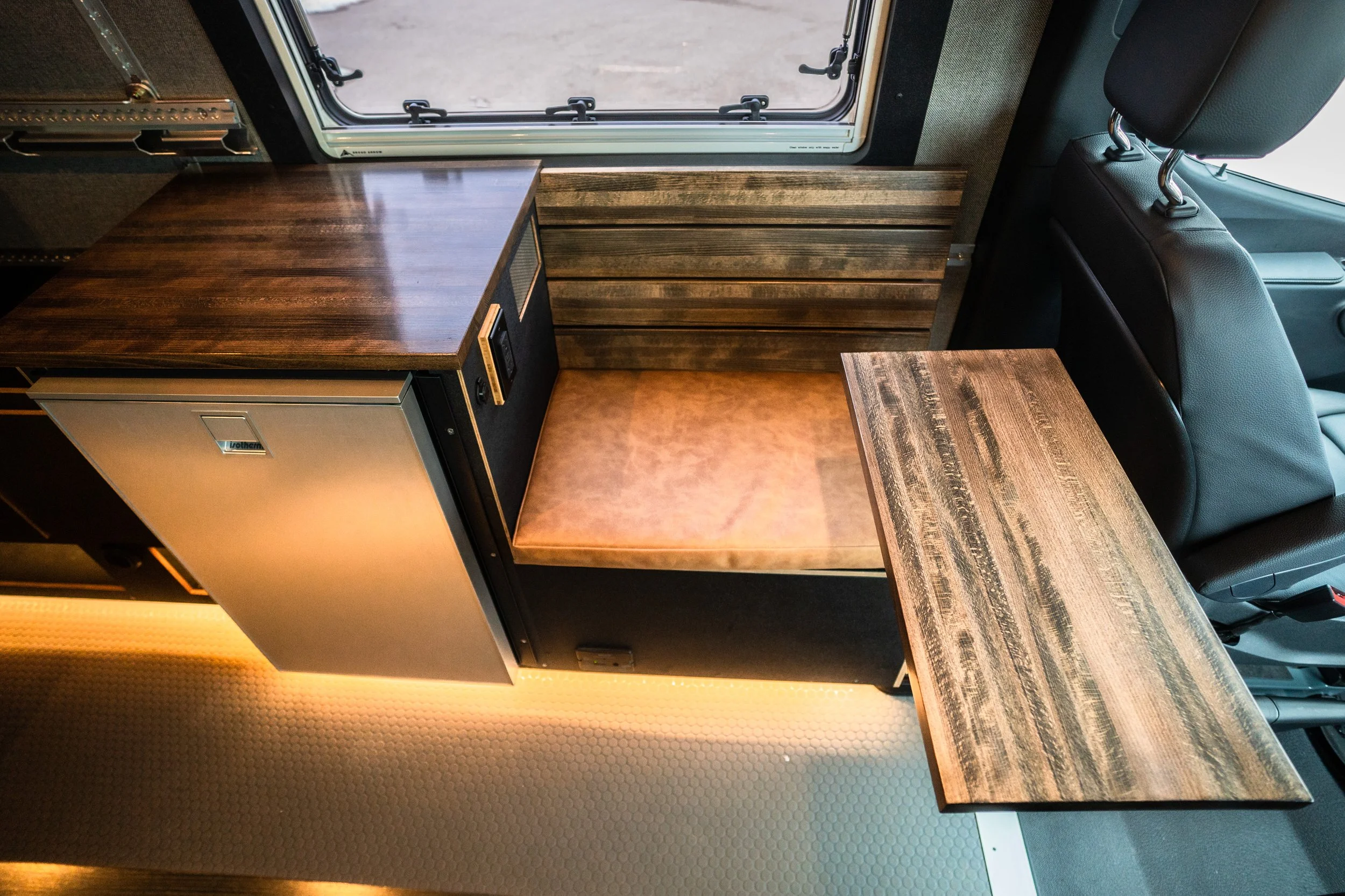 Interior of a camper van with a small refrigerator, a brown cushioned seat, a wooden table, and a window with a view of the outdoors.