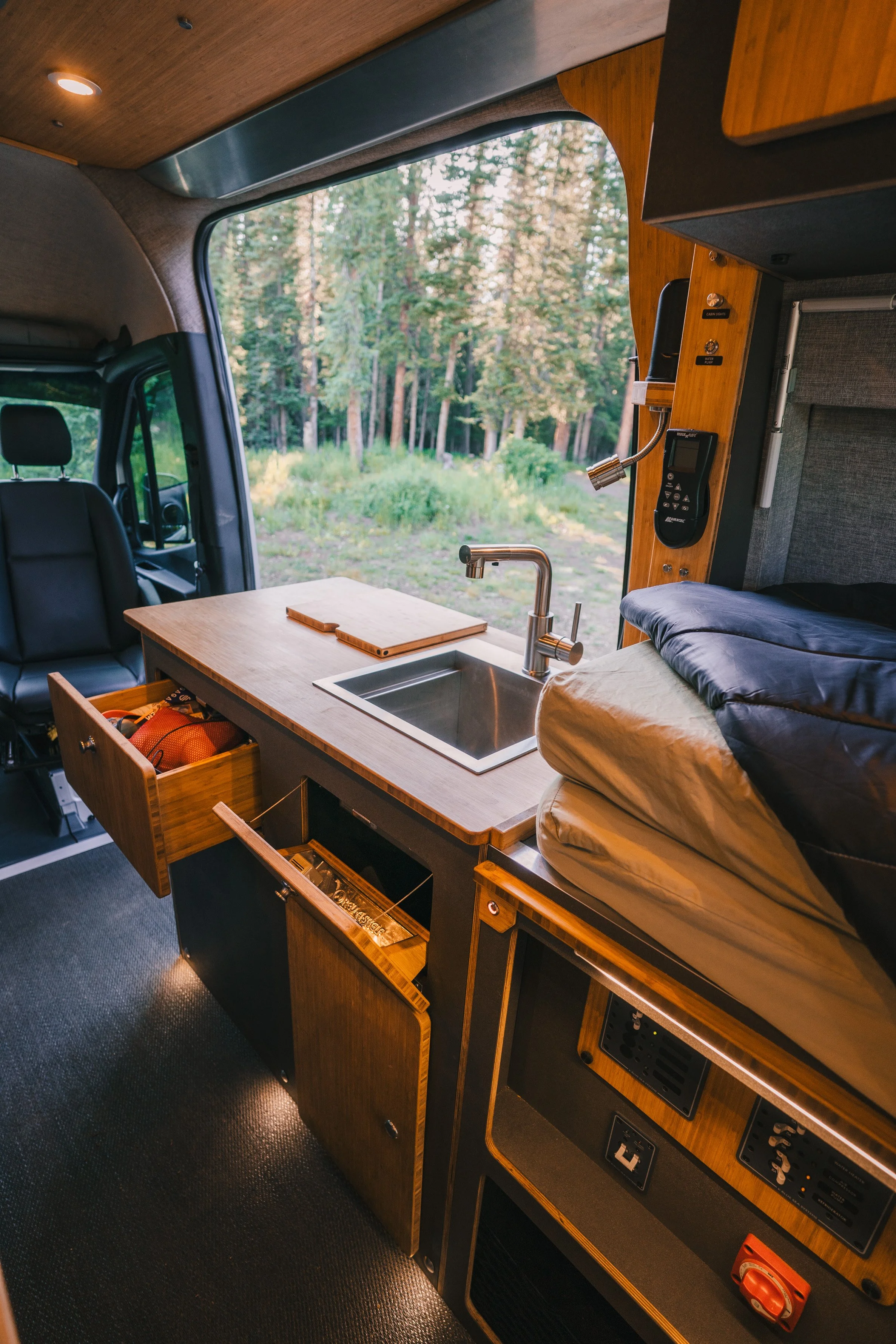 Interior of a camper van showing a compact kitchen with a sink, wooden cabinets, a bed, and view of trees outside through the window.