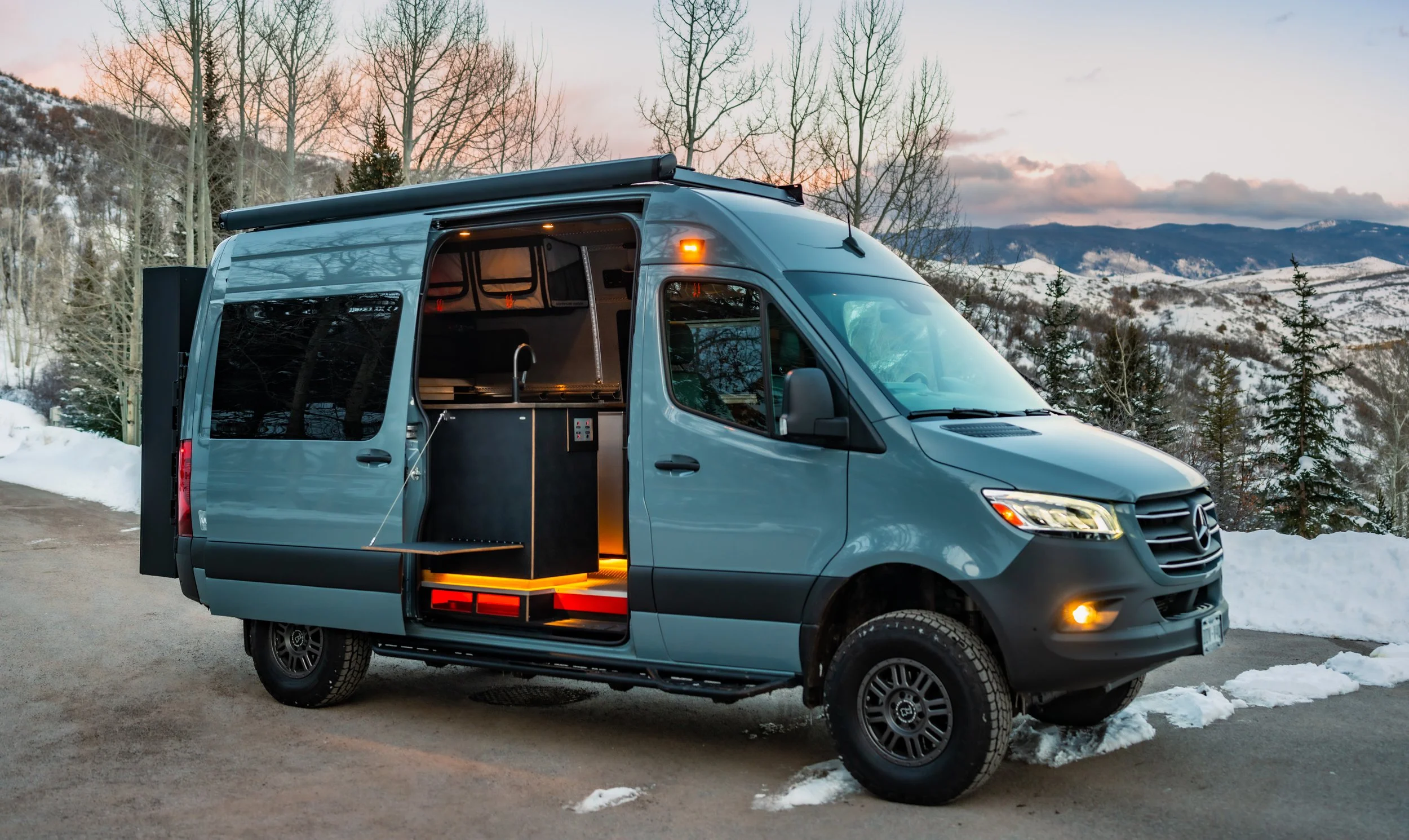 A blue Mercedes-Benz Sprinter van modified as a camper with an open side door showing a kitchenette, parked on a snowy mountain road during sunset with snow-covered mountains and trees in the background.