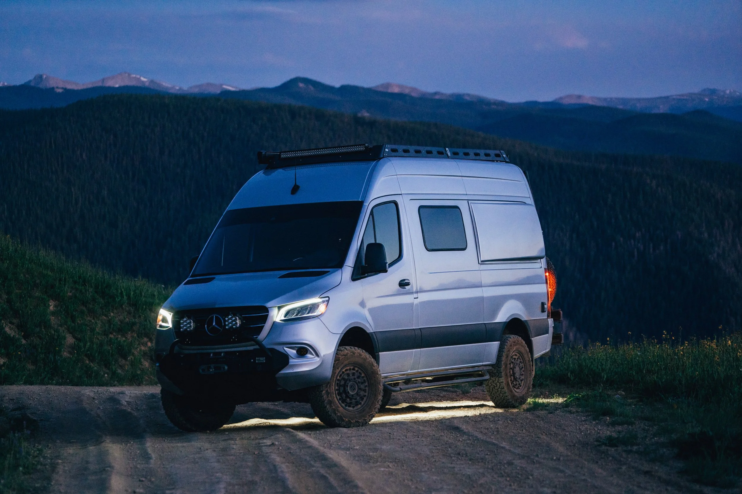 Silver camper van parked on a dirt trail with mountains and forest in the background during dusk.