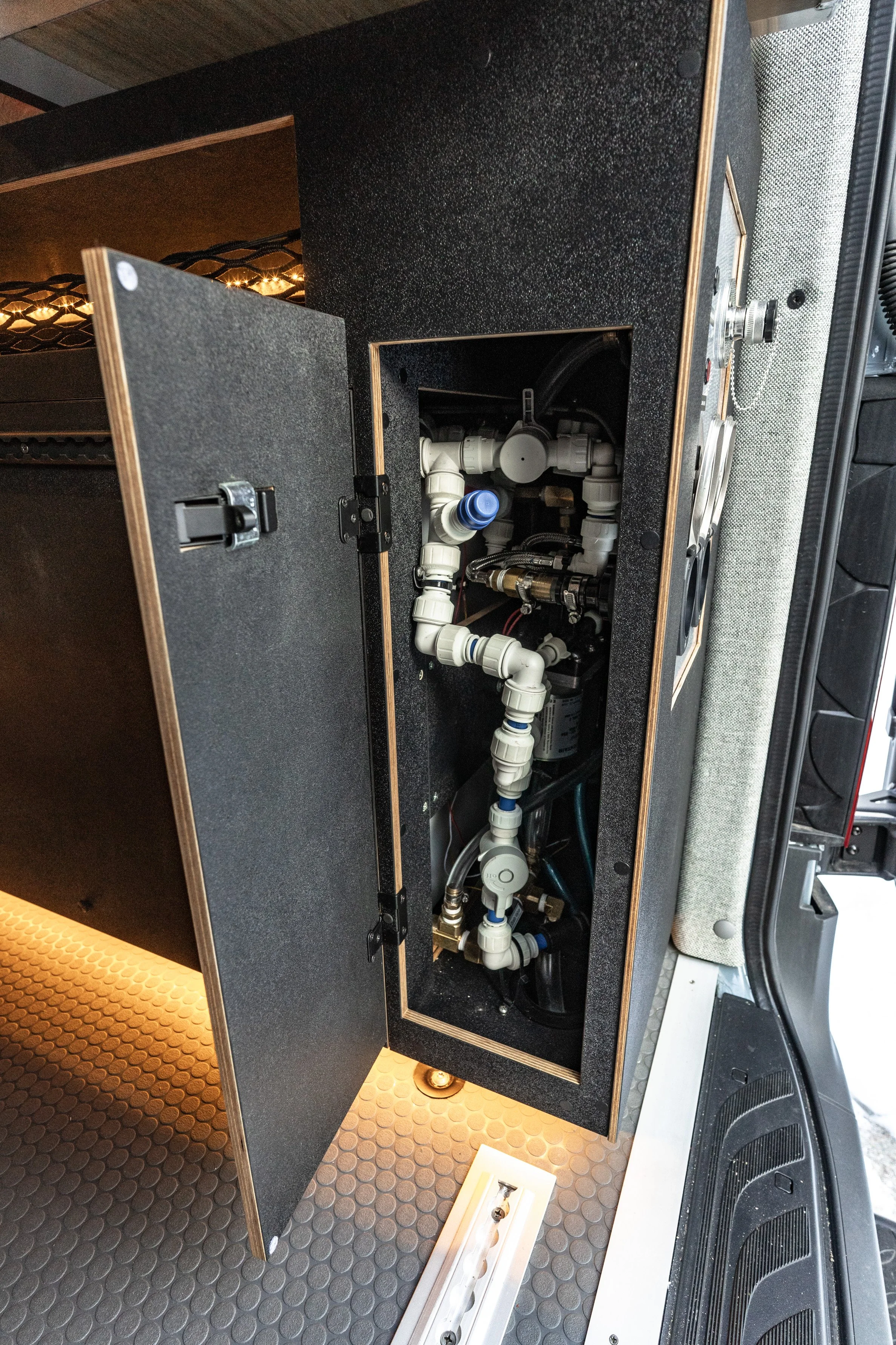Access panel open revealing plumbing pipes and fittings inside a cabinet in a vehicle or mobile unit.