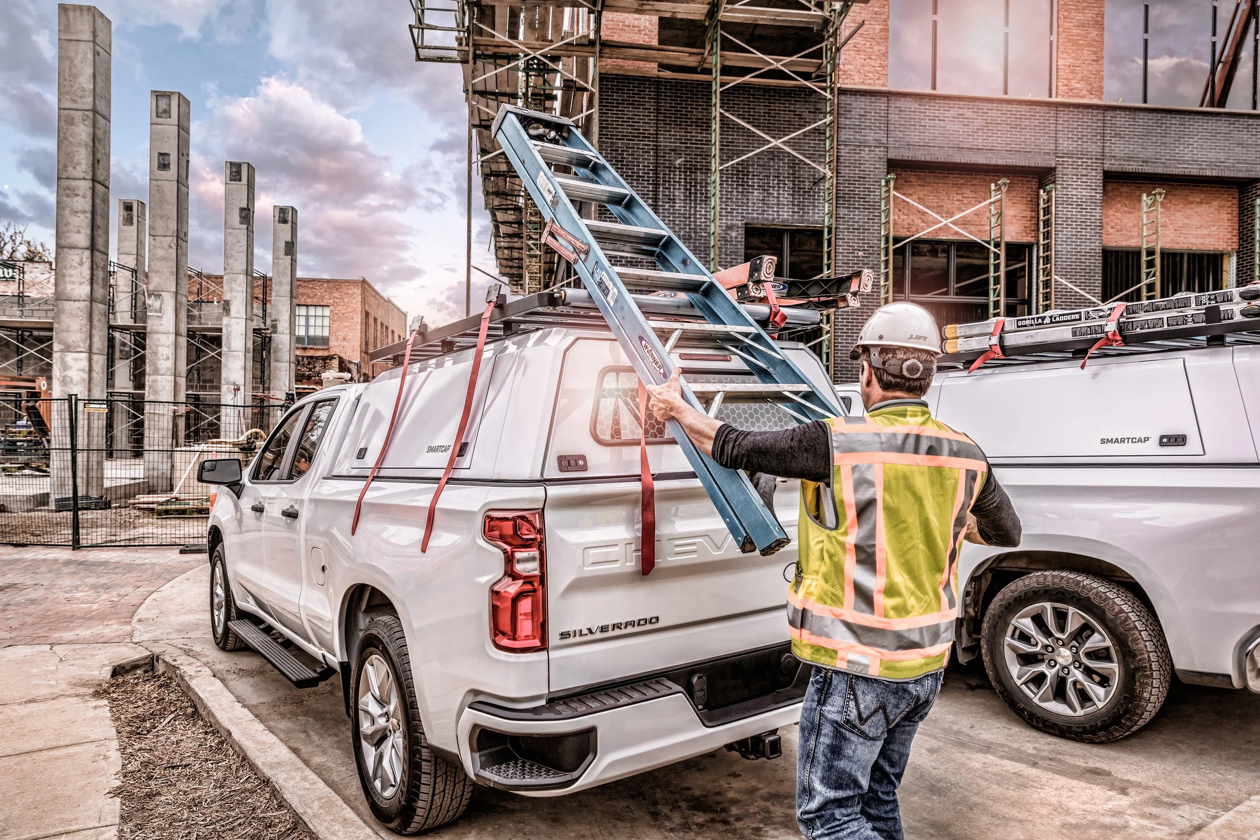 A construction worker wearing a safety helmet and vest is placing a ladder onto a white pickup truck at a construction site with buildings and scaffolding in the background.