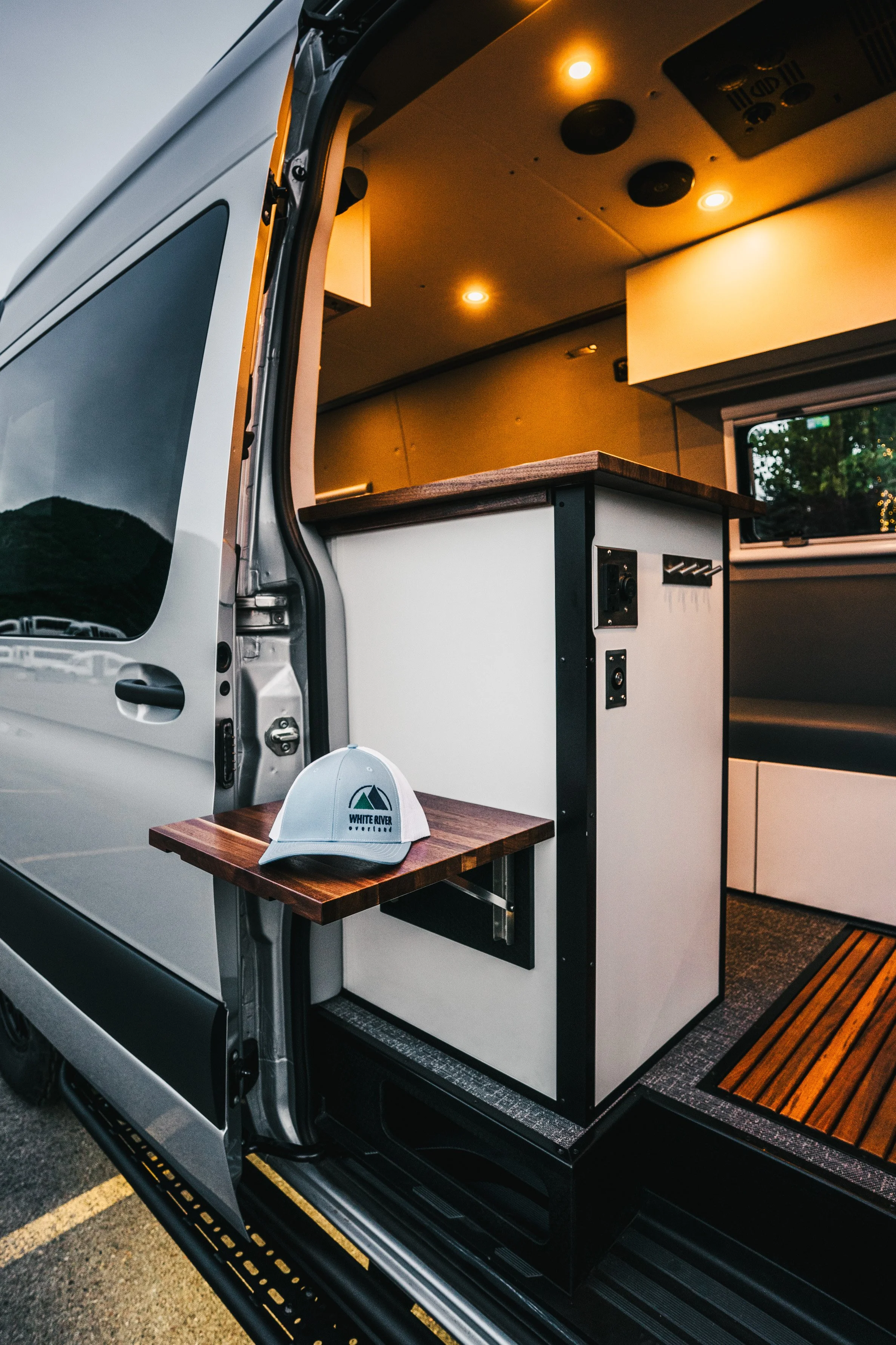 Interior of a van converted into a camper with a small folding table, white cabinetry, a window, and warm interior lighting.