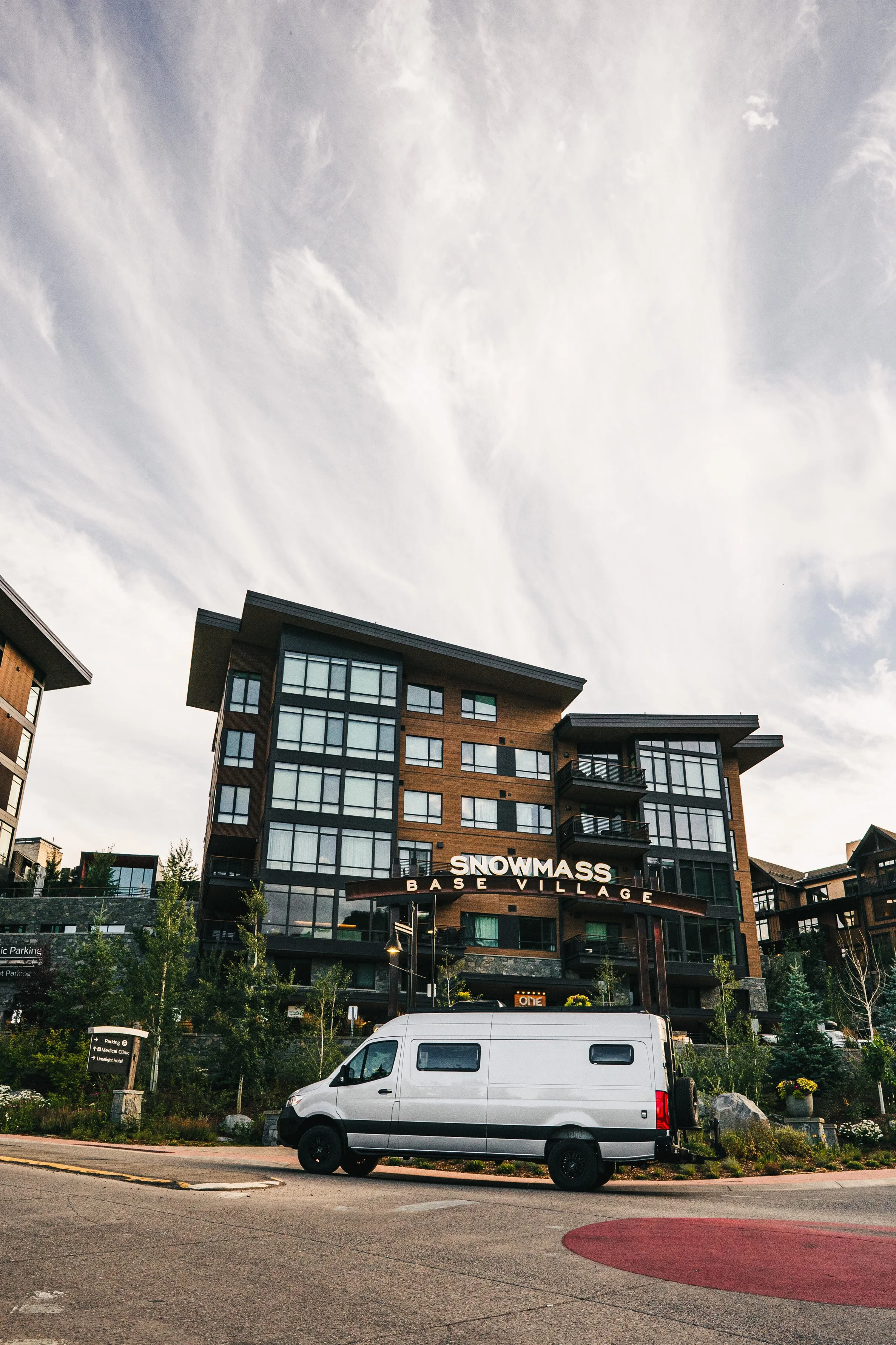 Multi-story residential building with the sign 'SNOWMASS BASE VILLAGE.' A silver Sprinter van is parked in front. The sky is partly cloudy.